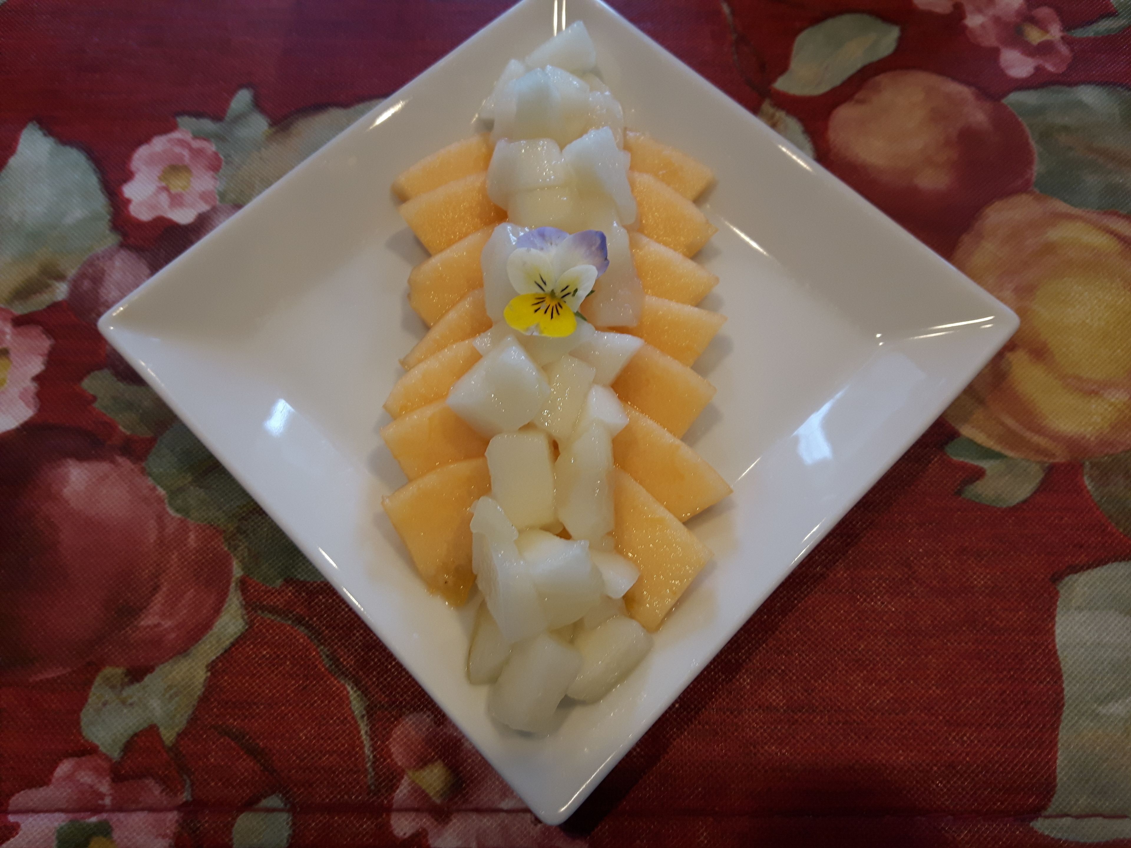 A white square plate with cubed melon and kiwi slices, placed on a table with a floral tablecloth.