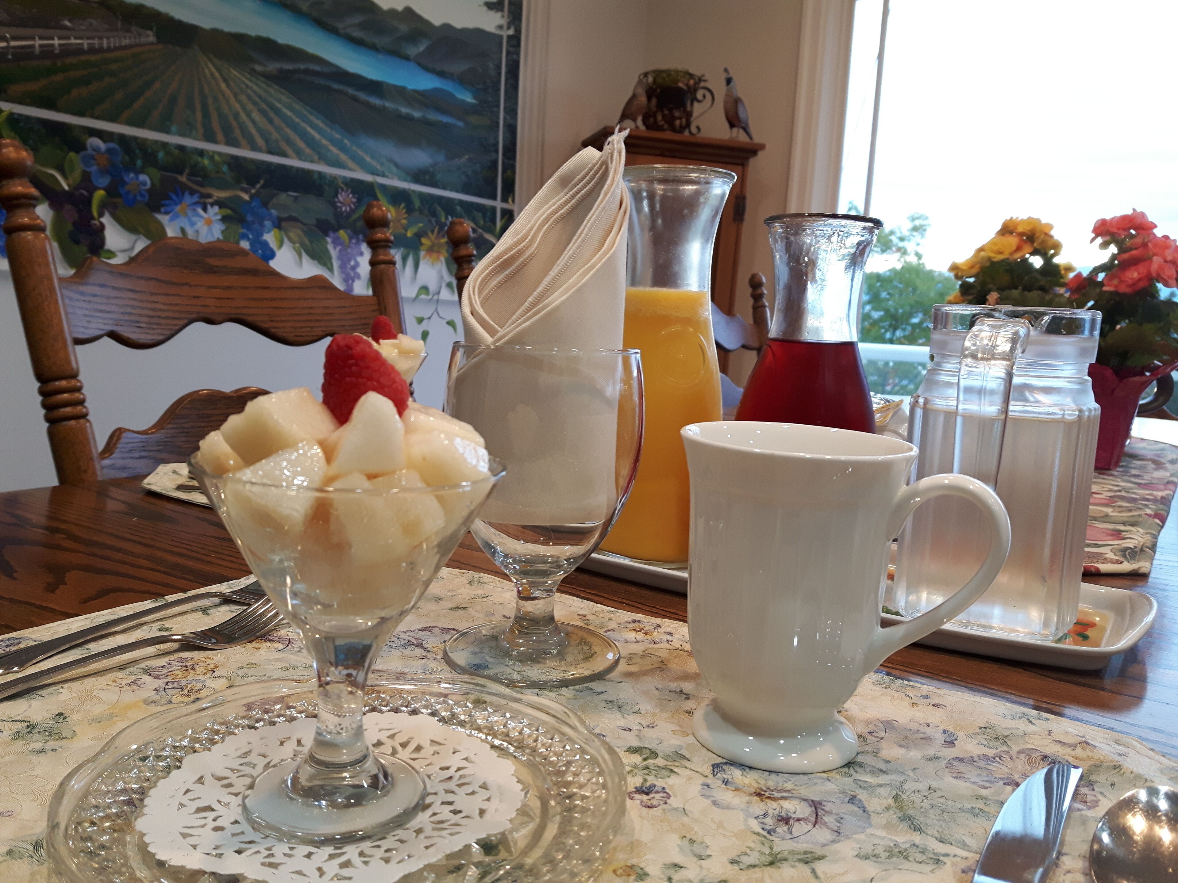 Un tavolo da pranzo con una tovaglia floreale, una ciotola di vetro piena di frutta, una brocca di succo d'arancia, una tazza bianca e un thermos.