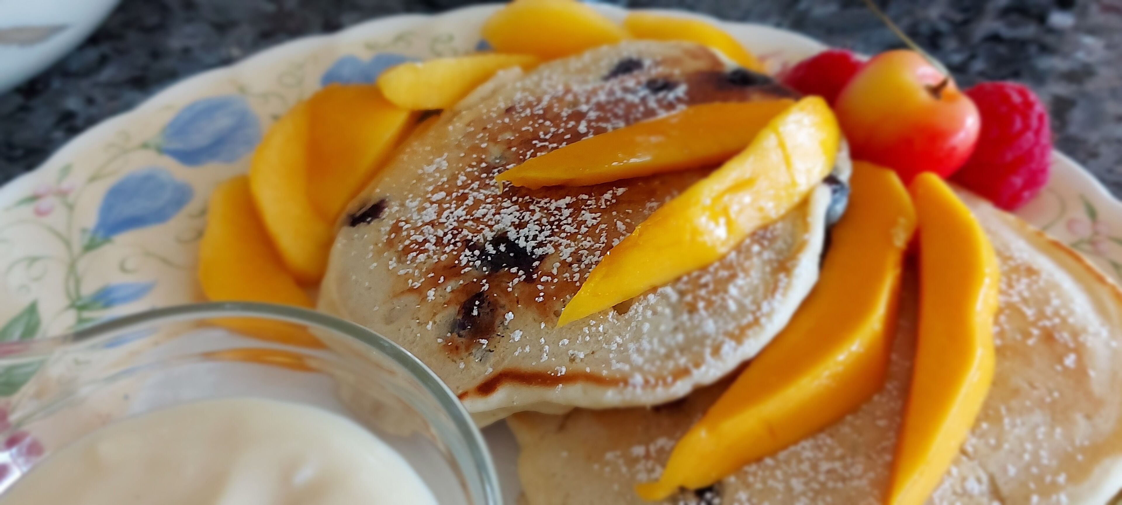 Un plato con panqueques cubiertos de mango en rodajas, una cereza y frambuesas.