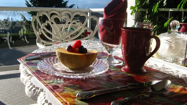 A table set with a bowl of fruit, a red cup, and a glass on a balcony overlooking a mountainous landscape. - A View to Remember Bed & Breakfast (West Kelowna)