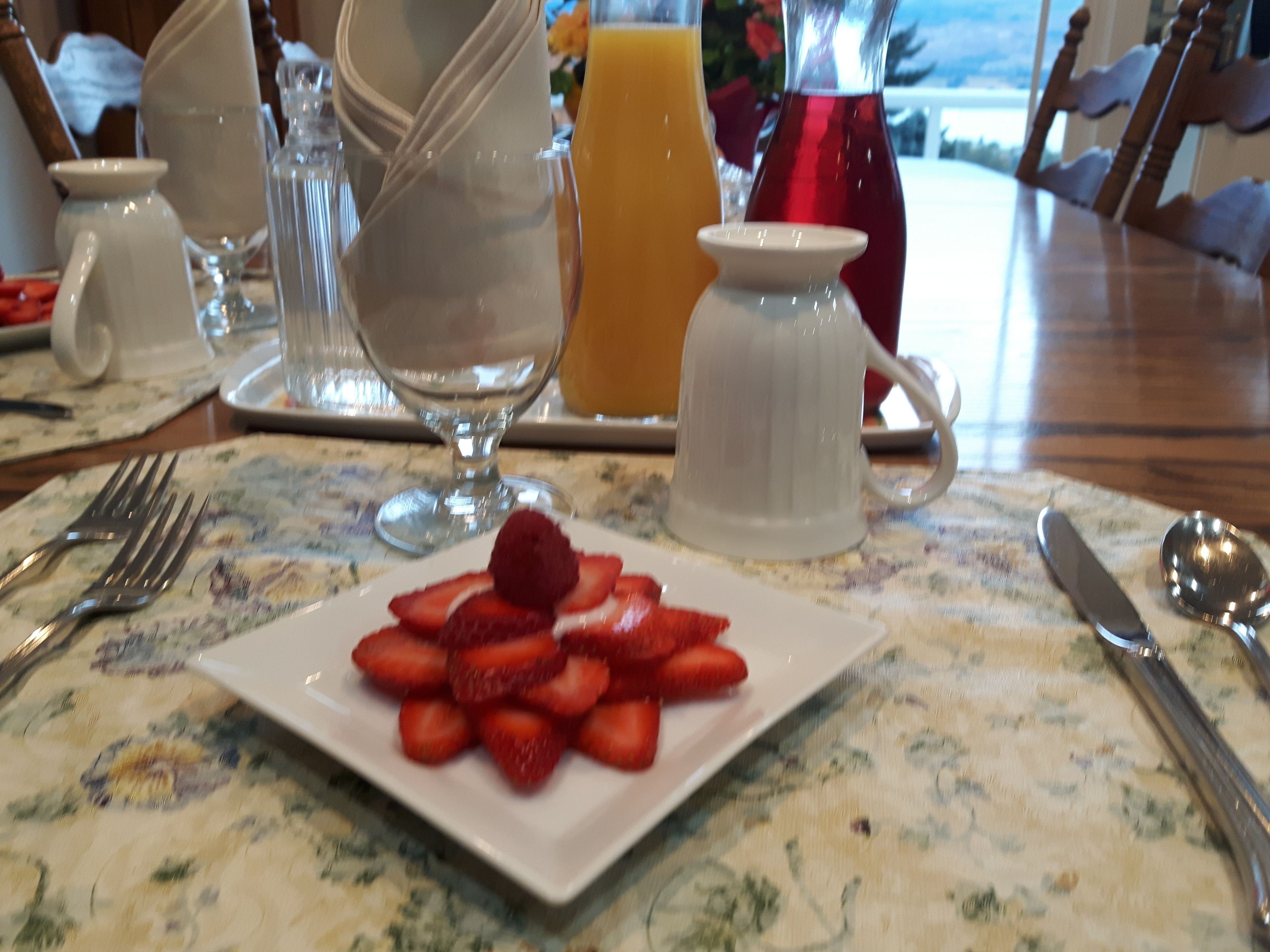 Una mesa de desayuno con un plato de fresas, una jarra de vidrio, un vaso de jugo de naranja y una jarrón blanco.