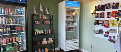 A vending machine stocked with snacks and beverages, a refrigerator with a 'DASANI' sign, and a wall displaying various packaged goods.
