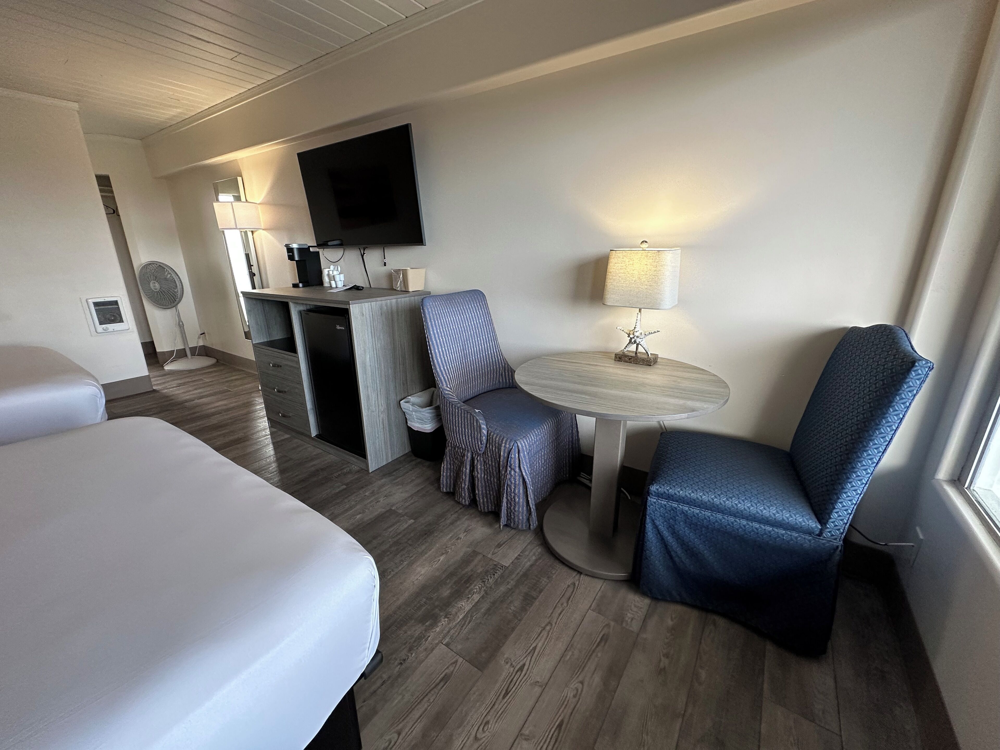 Habitación de hotel con dos camas, un escritorio con televisor de pantalla plana, una kitchenette pequeña, una mesa redonda con lámpara y una ventana.