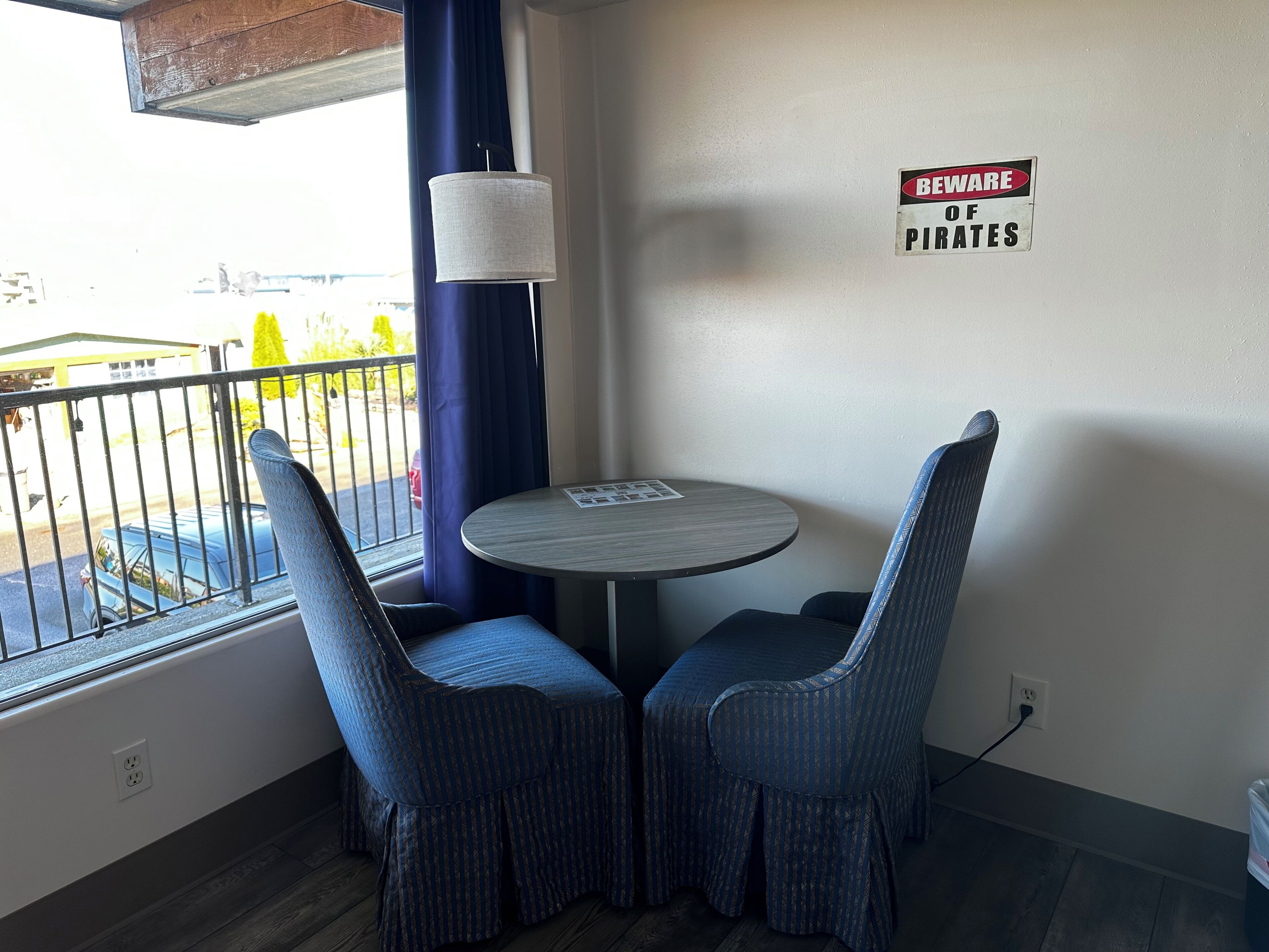 A corner room with two upholstered chairs, a round table, a lamp, a 'Beware of Pirates' sign, and a window overlooking a parking area.