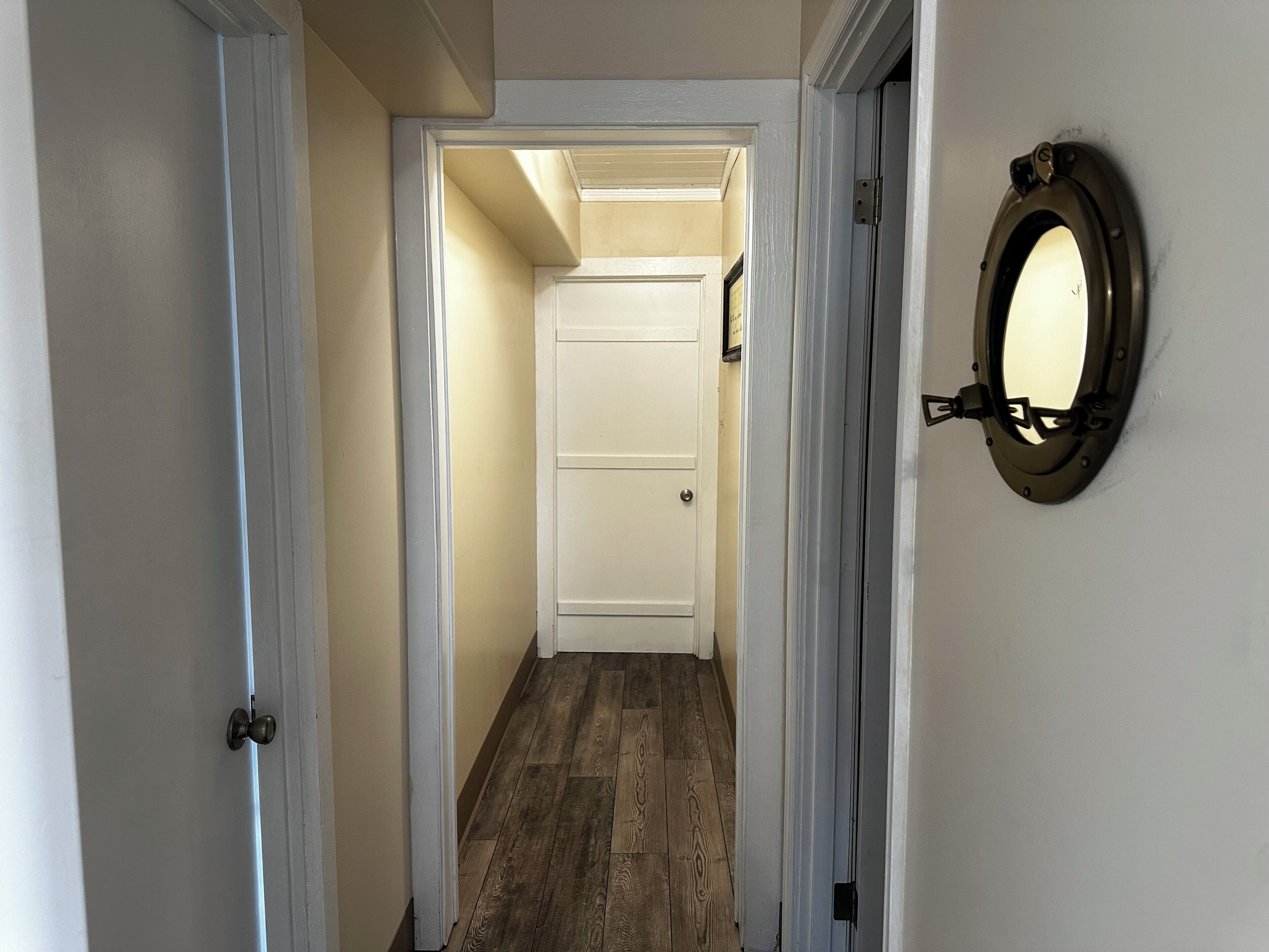 A narrow hallway with wooden flooring, white walls, and a white door at the end.
