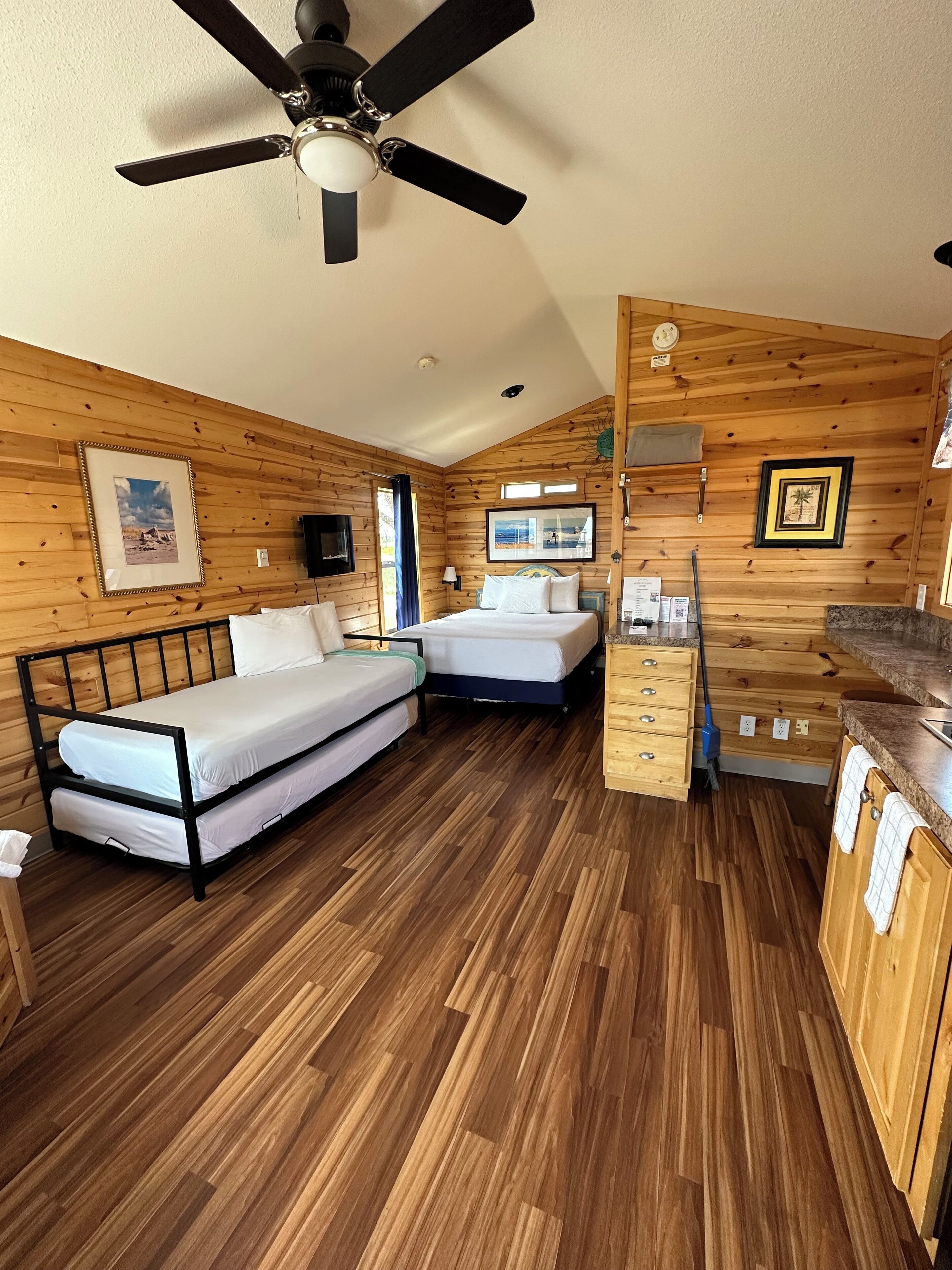 Una habitación tipo cabaña compacta con dos camas, ventilador de techo, paredes de madera, kitchenette pequeña y suelo de madera.