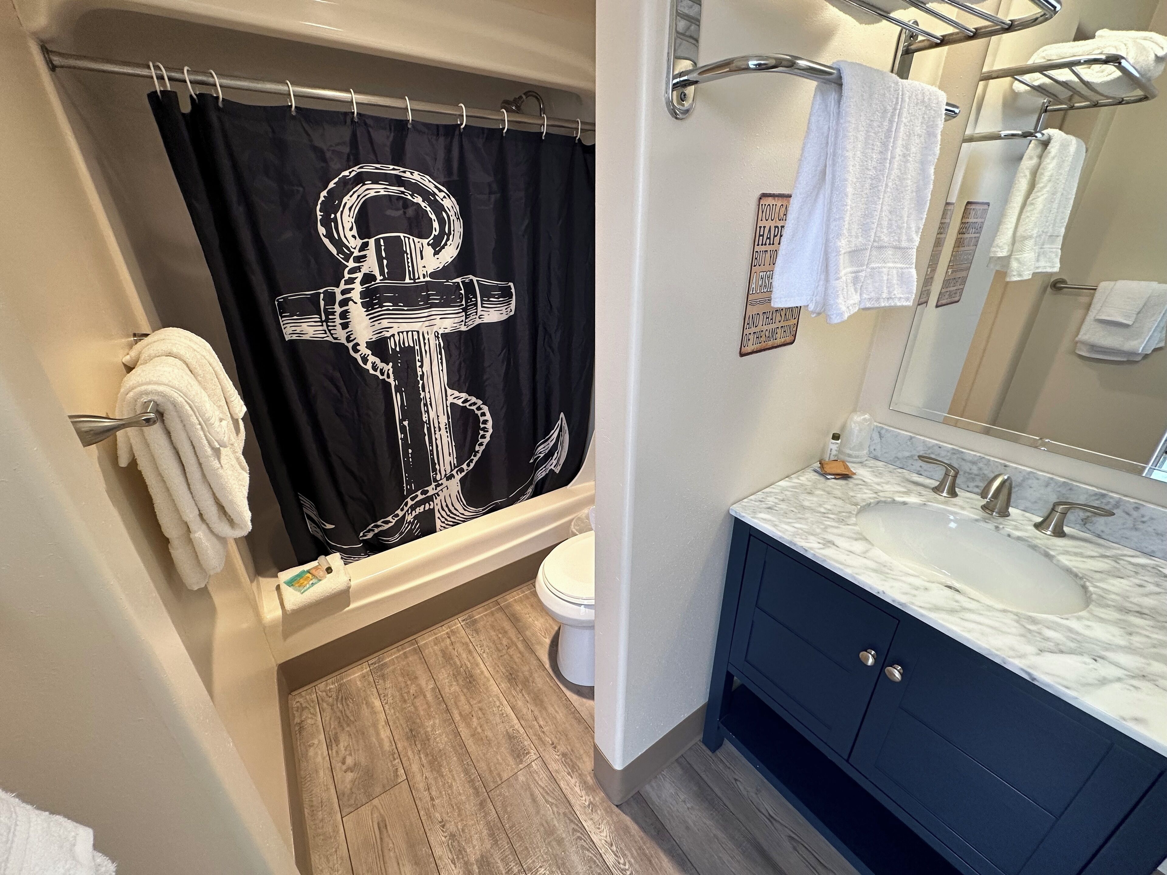 Um banheiro com cortina de chuveiro estampada com âncora em preto e branco, vaso sanitário branco, pia com bancada de mármore e armário escuro azul.