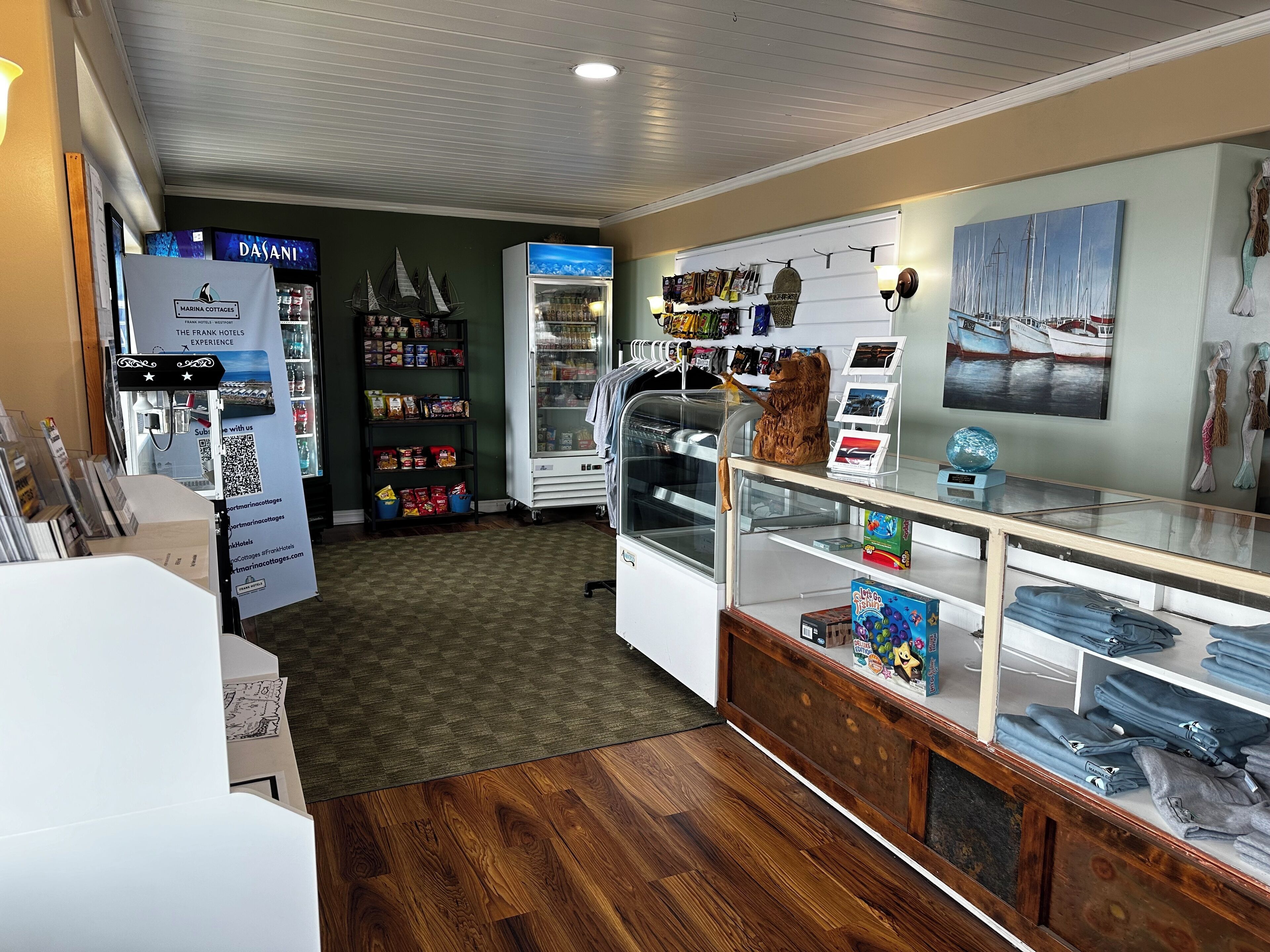 Interior de agência de viagens com chão de madeira, vitrine de vidro com folhetos, um refrigerador e parede com arte decorada com tema náutico.