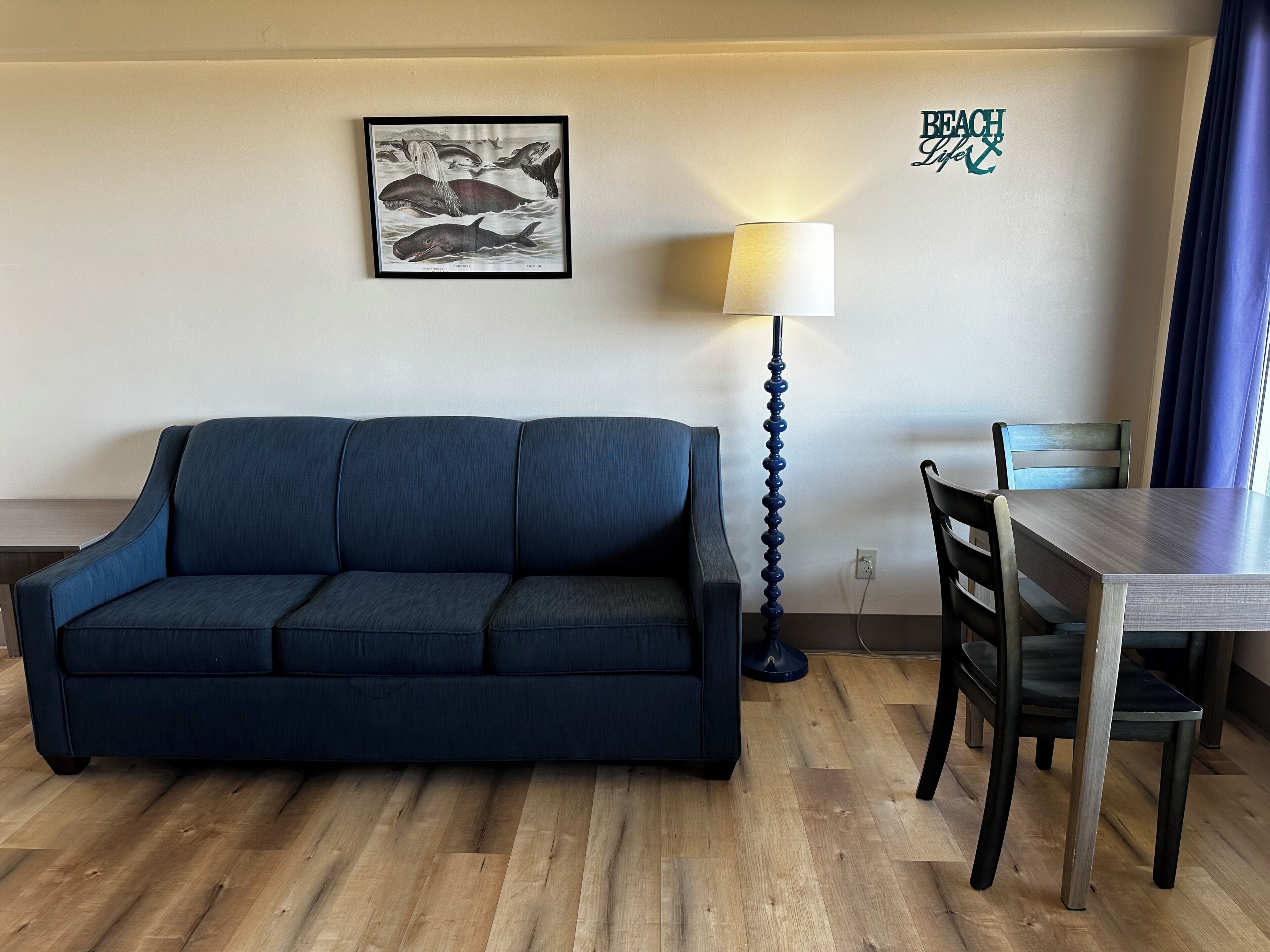 Ein Zimmer mit einem dunkelblauen Sofa, einem Holzesstisch mit Stühlen, einer Lampe und einem gerahmten Bild von Walen an der Wand.