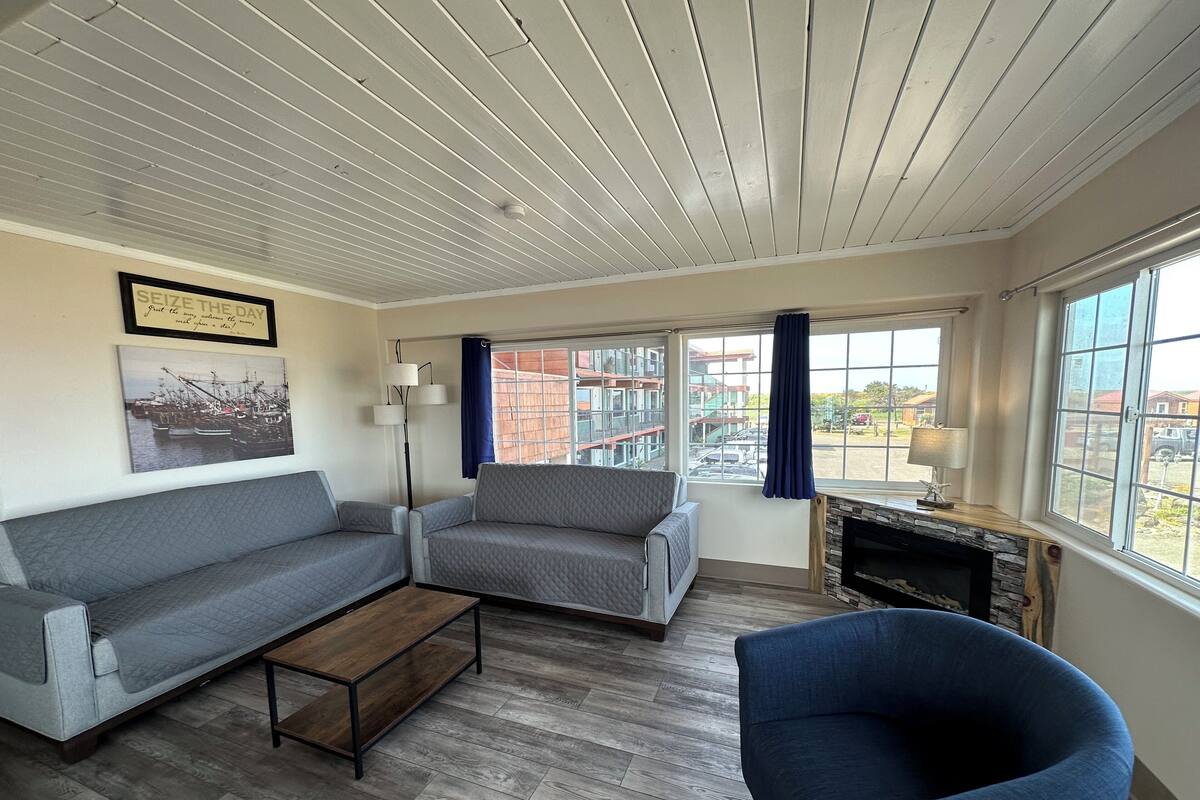 Sala de estar com sofá cinza, poltrona azul, mesa de centro de madeira e lareira. Há grandes janelas com cortinas, um quadro na parede e teto com vigas de madeira.