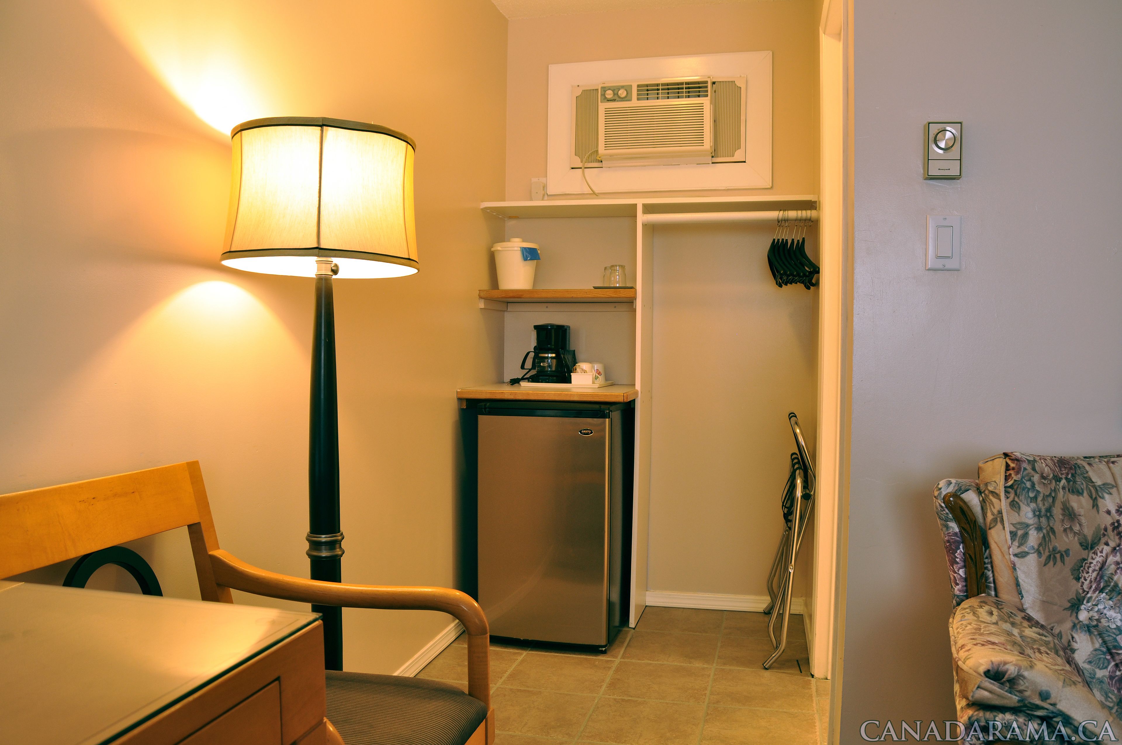 A compact hotel room with a desk, chair, lamp, mini-fridge, coffee maker, and a closet with hanging clothes.