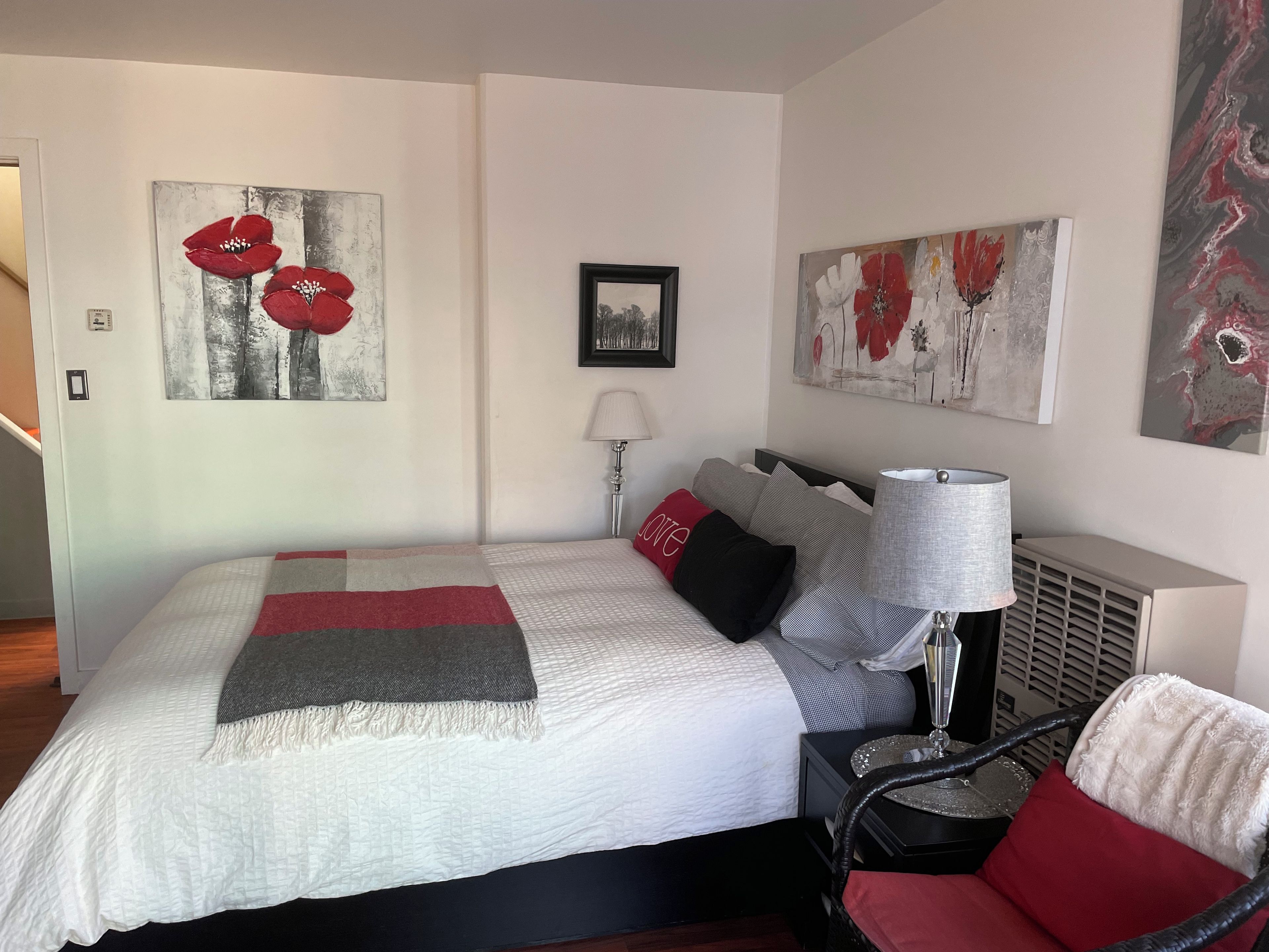 A neatly made bed with a white duvet, red and black pillows, and a grey throw. There are two wall-mounted lamps and a framed picture above the bed.
