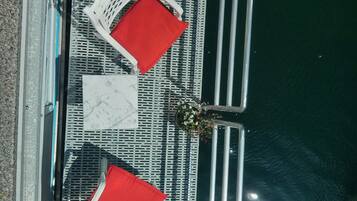 A boat deck with two white wicker chairs with red cushions, a white marble table, and a metal railing.