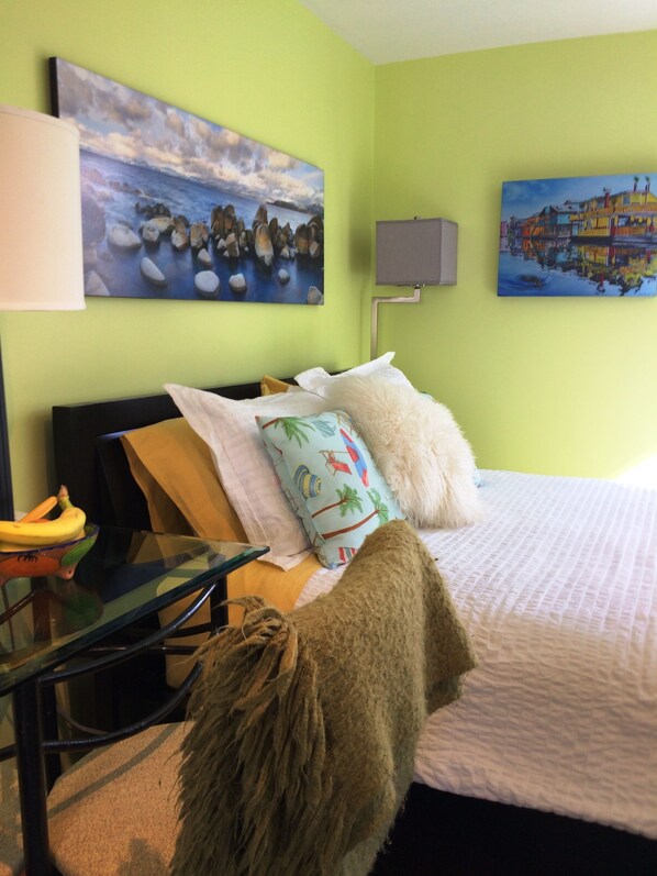 Una habitación con cama, almohadas, una manta, una mesa con un plátano, una lámpara y dos cuadros en la pared.