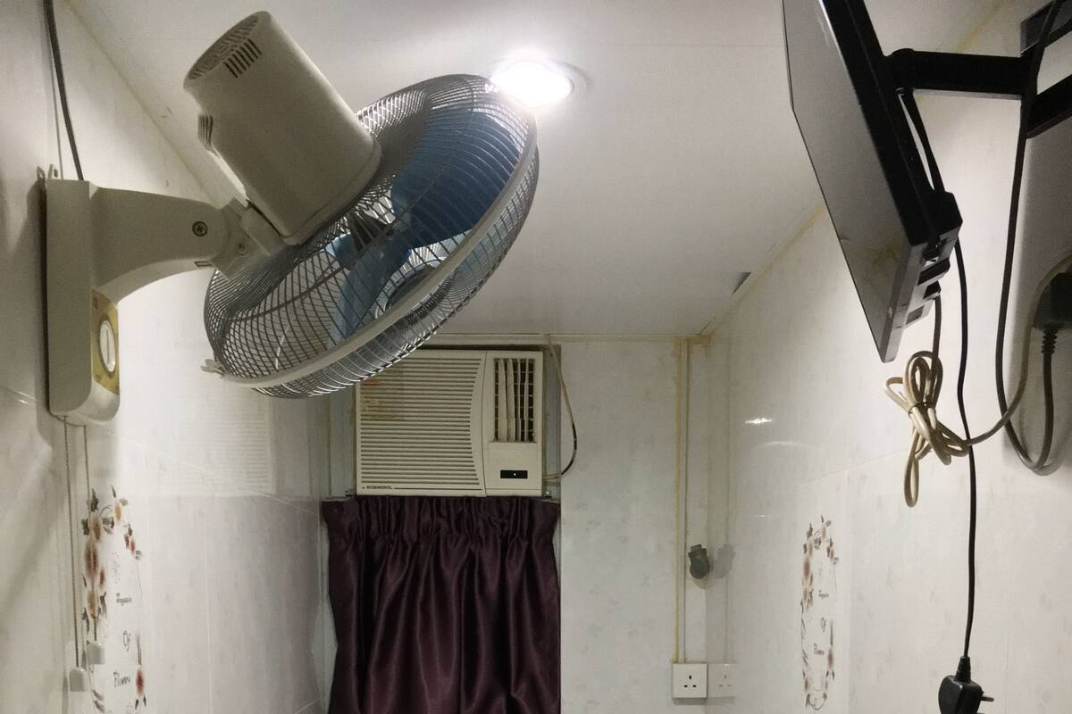 A small, single-bed room with a headboard, a wall-mounted air conditioner, a ceiling fan, and a power outlet.