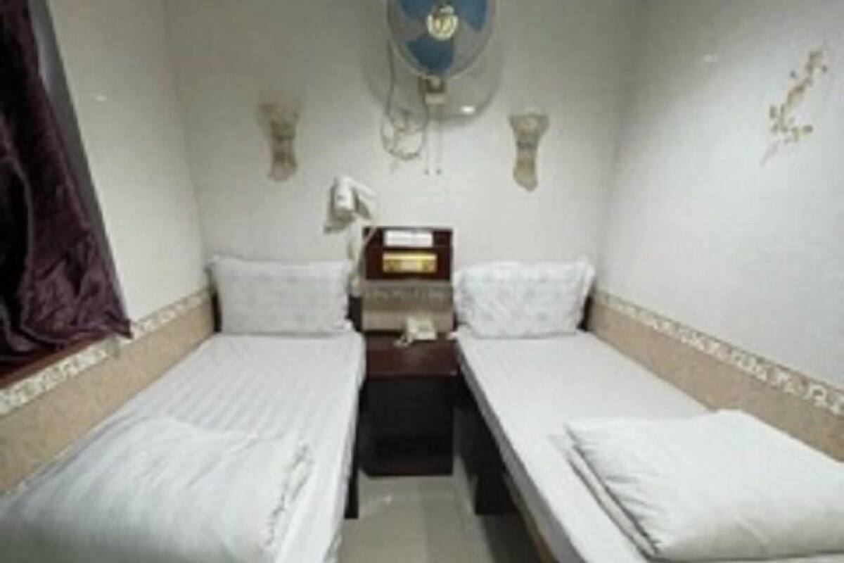 A room with two single beds, a bedside table with a lamp, a wall-mounted fan, and a small decorative item on the wall.