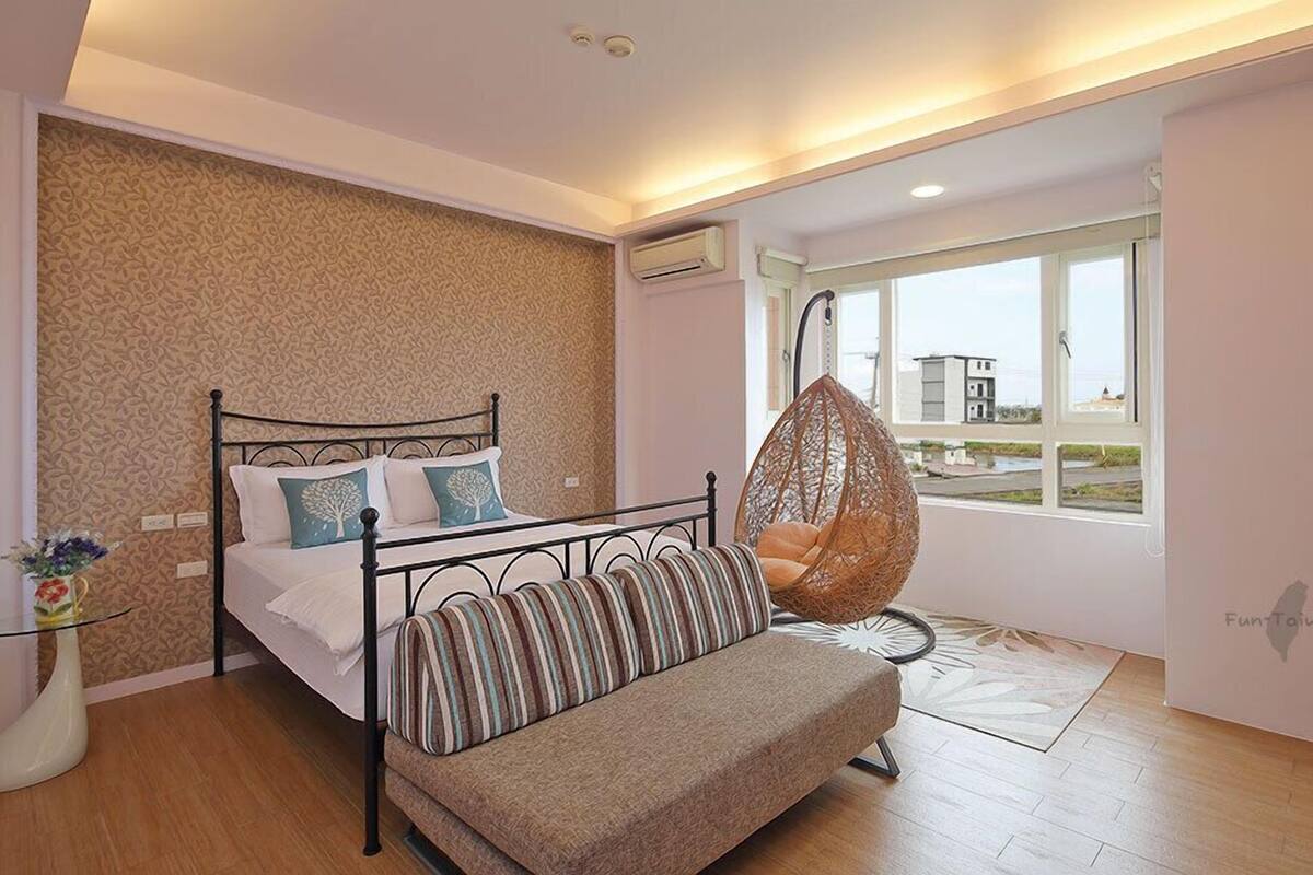 Een slaapkamer met een bed, een hangstoel, een bank, een vaas met bloemen en een raam met uitzicht.