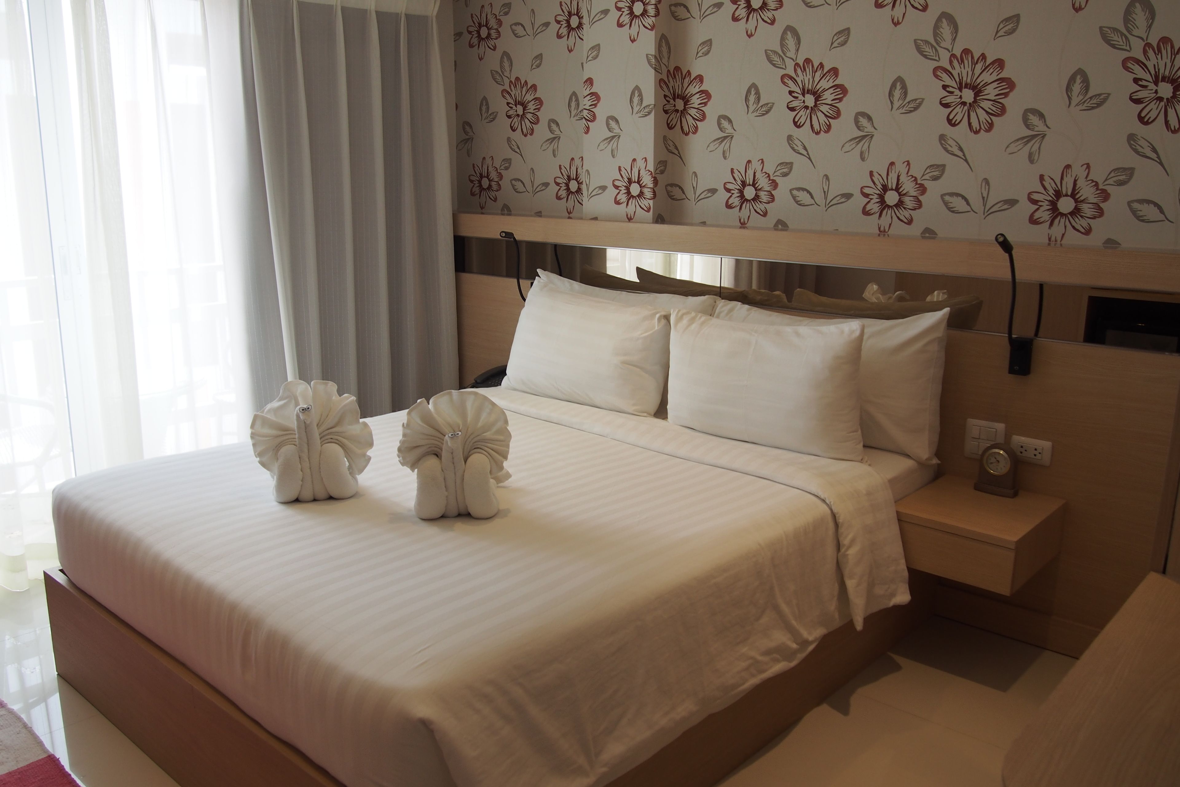A hotel room with a large bed, a nightstand, and a floral-patterned wall.
