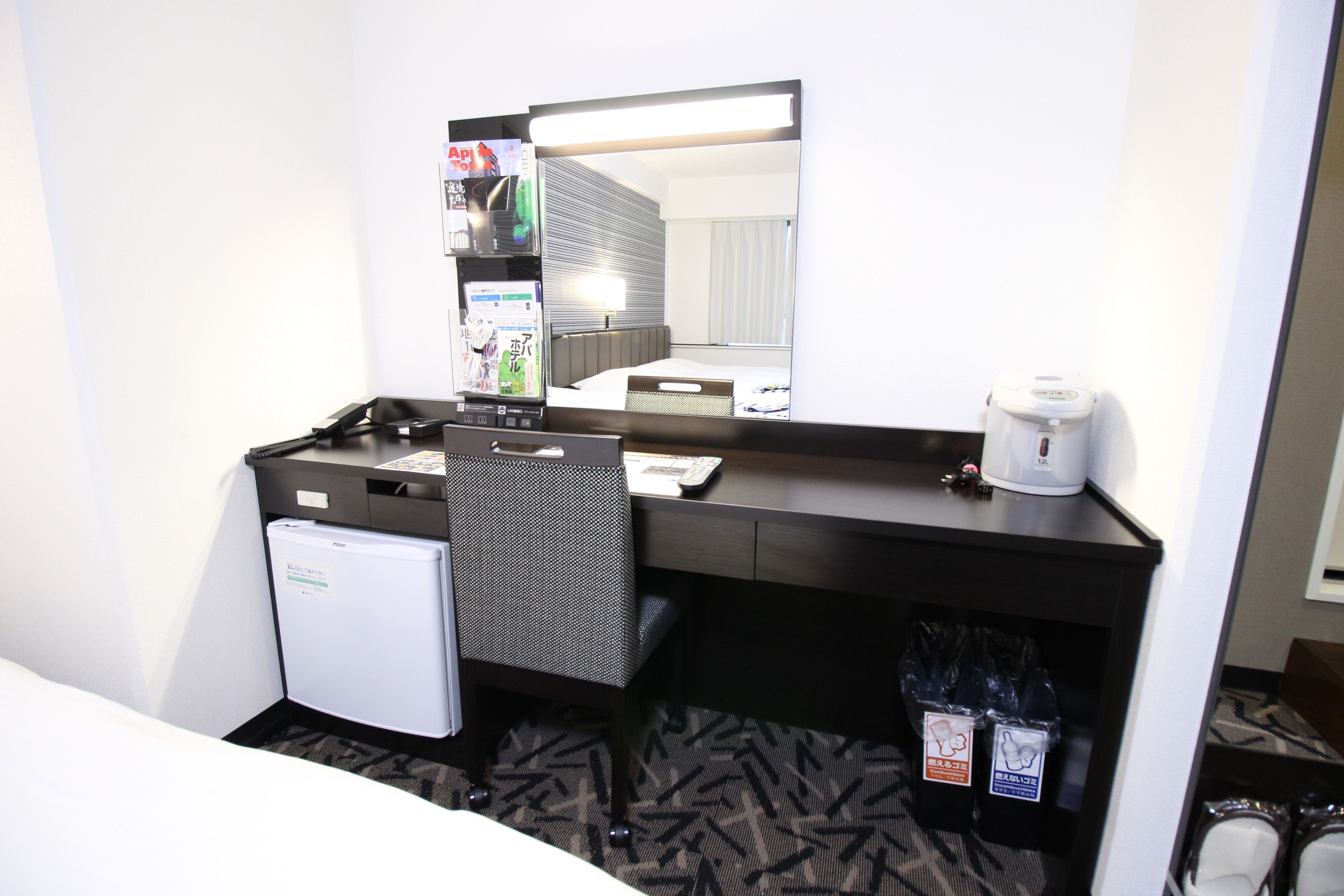 A hotel room with a small refrigerator, a desk with a chair, a coffee maker, and a trash bin.