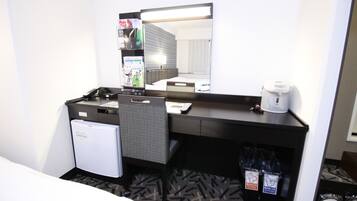 A hotel room with a small refrigerator, a desk with a chair, a coffee maker, and a trash bin.