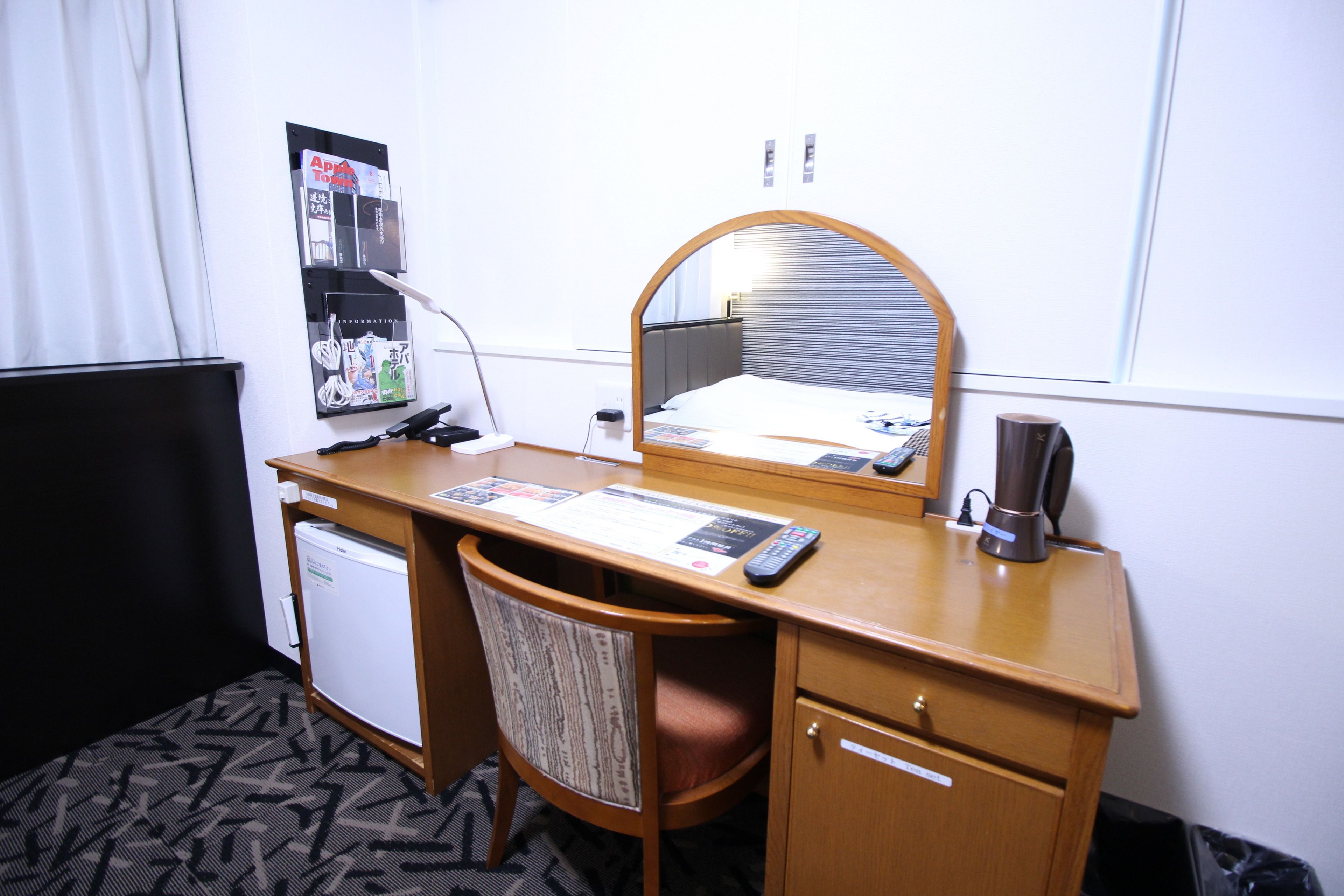 Une chambre d’hôtel avec un bureau en bois, une chaise, un petit réfrigérateur, un miroir et un panneau d’affichage où sont accrochés divers objets.