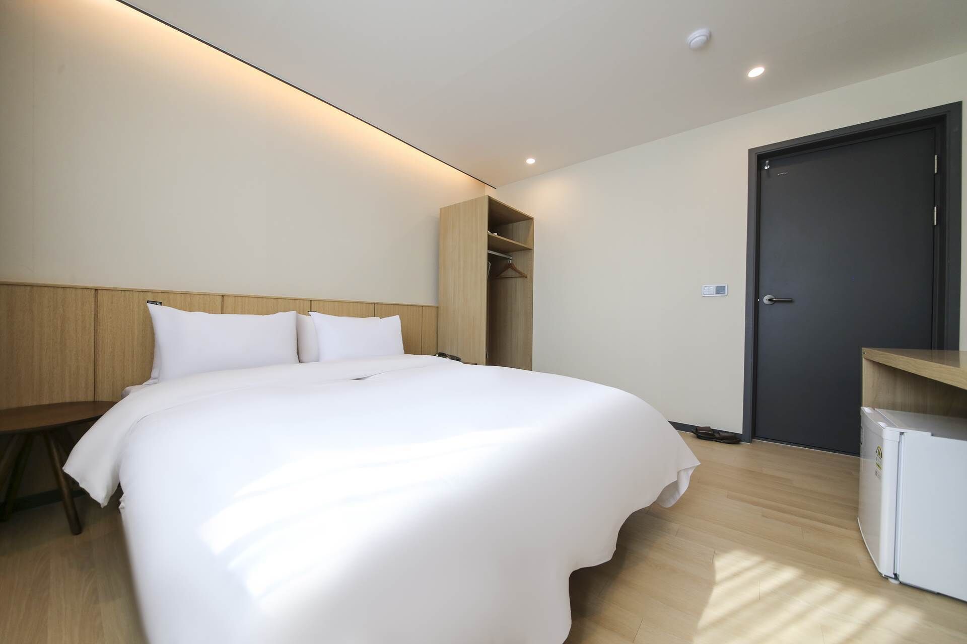 A modern hotel room with a large bed, wooden headboard, built-in closet, and a small refrigerator.