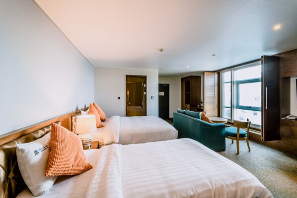 Habitación de hotel con dos camas, un sofá, una silla, un escritorio y un ventanal grande.