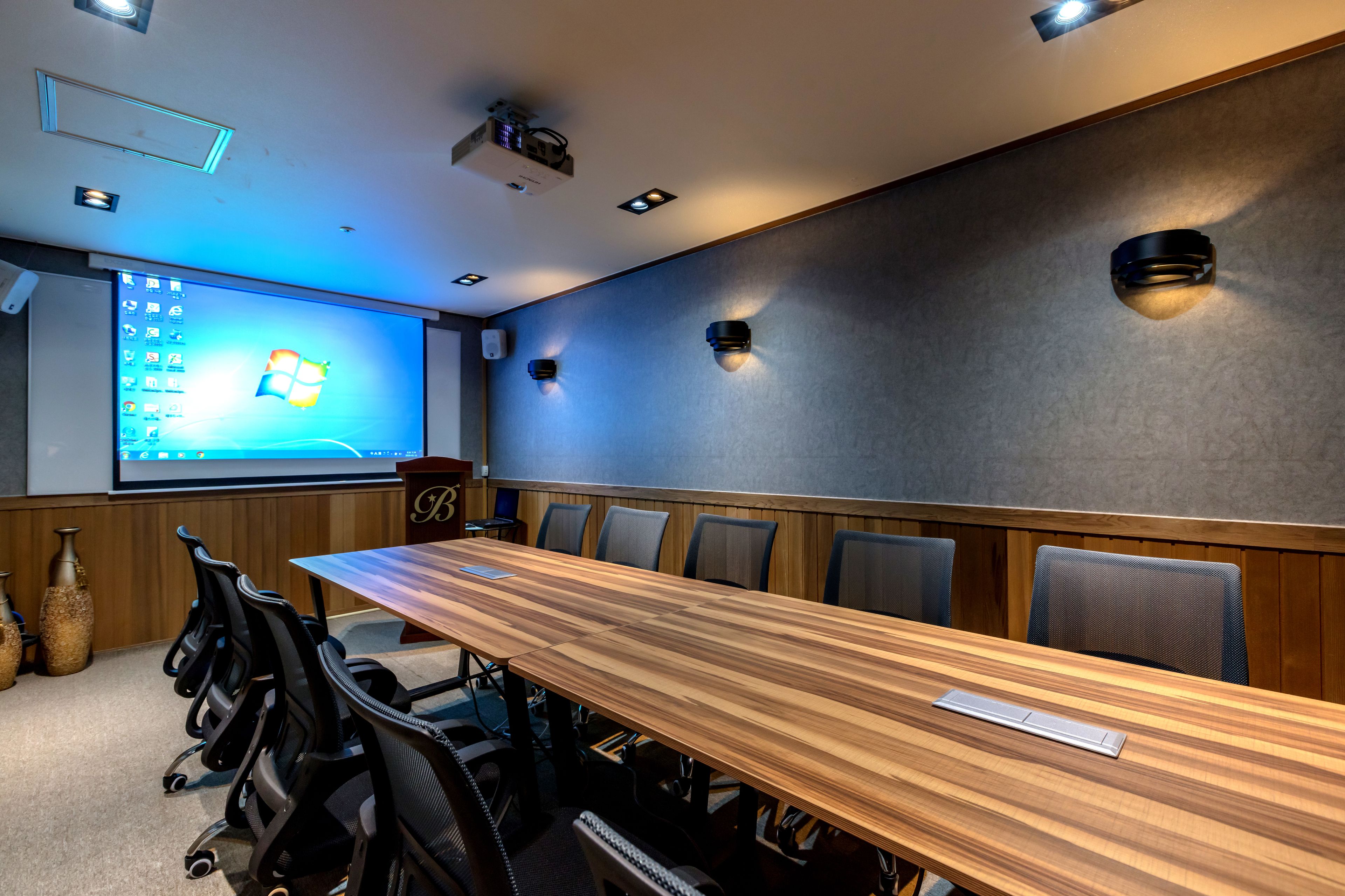Un salón de conferencias con una larga mesa de madera, sillas de oficina negras, una pantalla de proyector que muestra un escritorio de Windows y una pared oscura con tres luces empotradas.