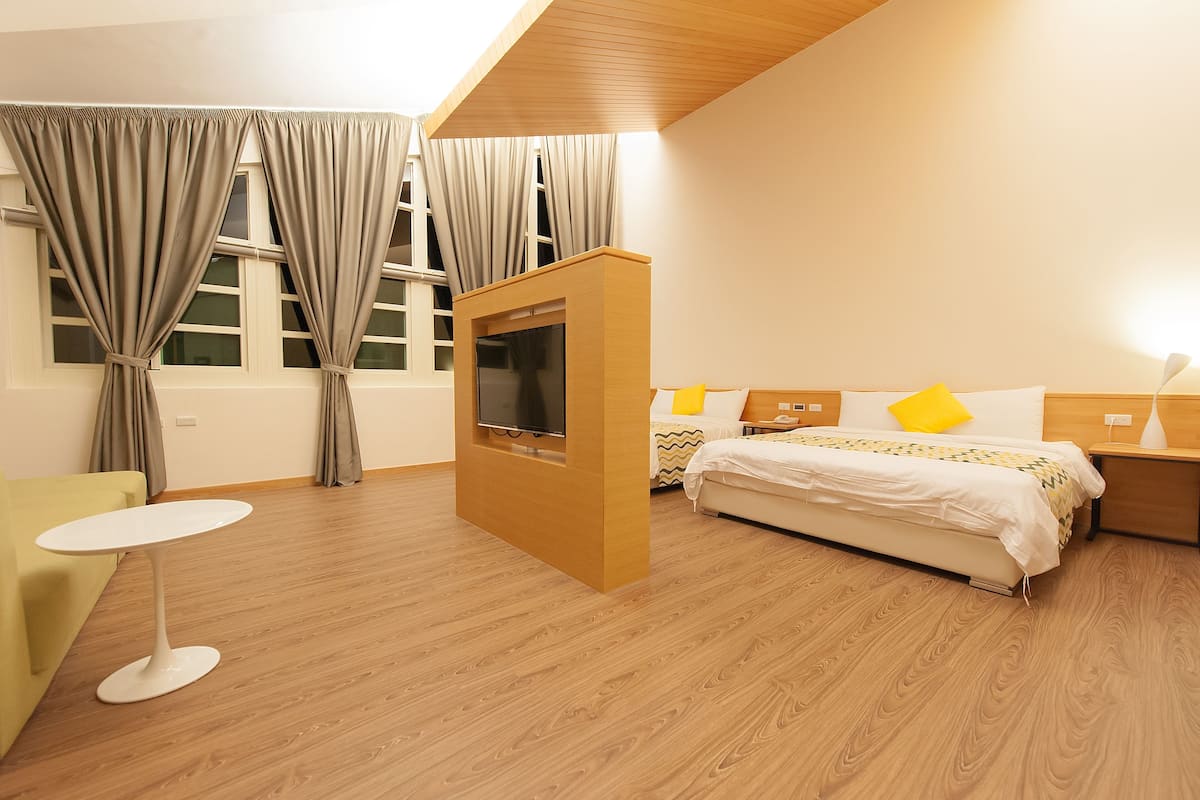 Habitación de hotel moderna con suelo de madera, una cama con ropa de cama blanca y almohada amarilla, una mesita redonda pequeña y un televisor montado en un mueble de madera.