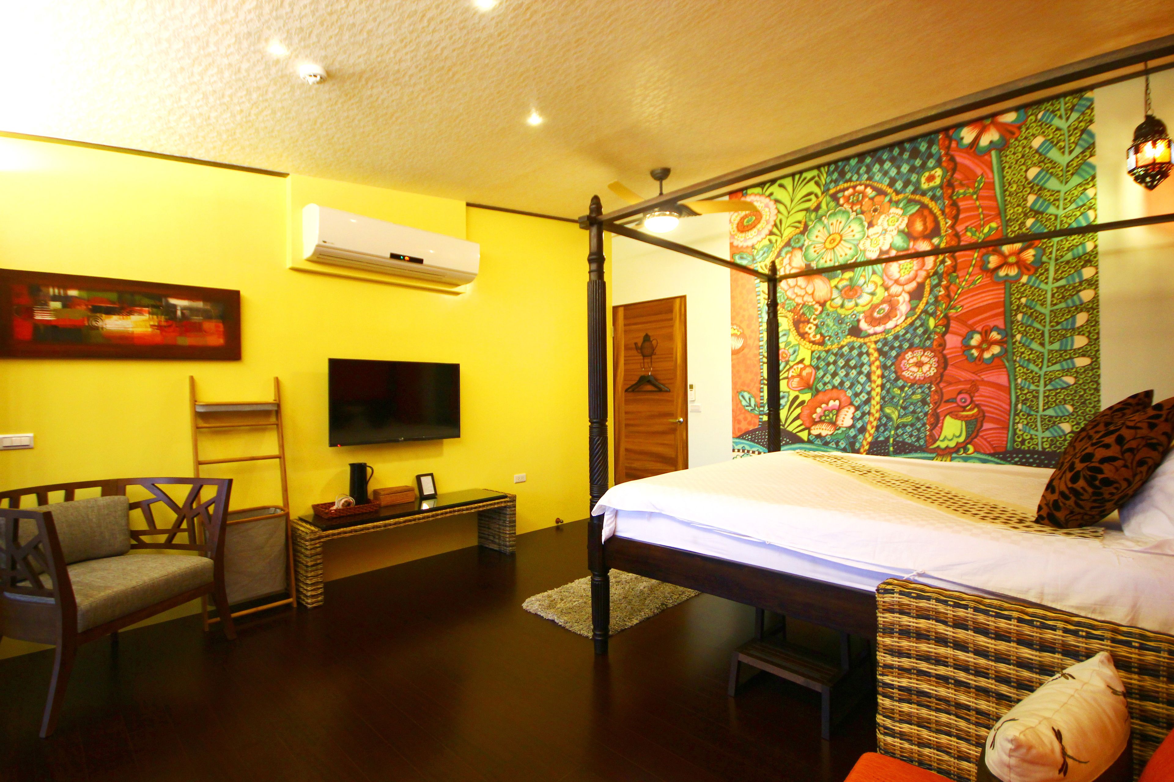 A bedroom with a bed, a television, a chair, and a colorful tapestry on the wall.