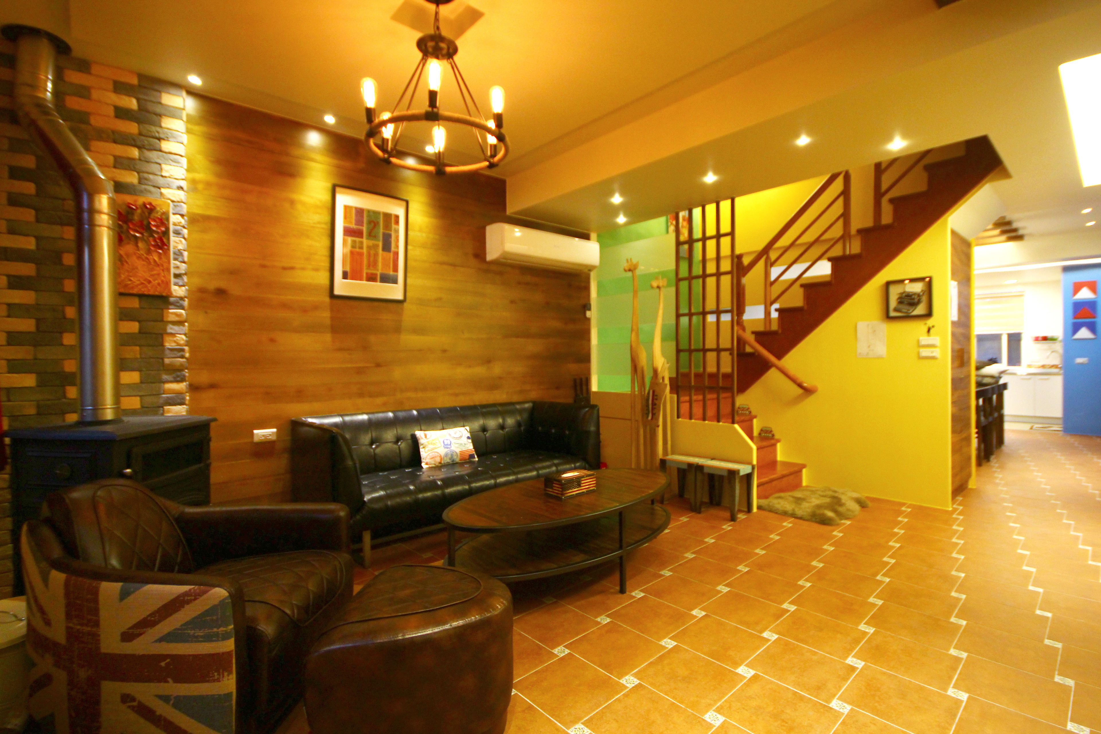 A modern living room with a leather sofa, a glass coffee table, and a patterned armchair. A staircase leads to an upper level, and the room features a wooden accent wall and a hanging light fixture.