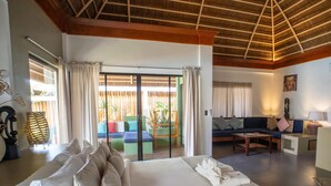 A hotel room with a large bed, a sofa, a dining table, and a view of a wooden deck and outdoor seating area. - Villa Kasadya (Panglao)