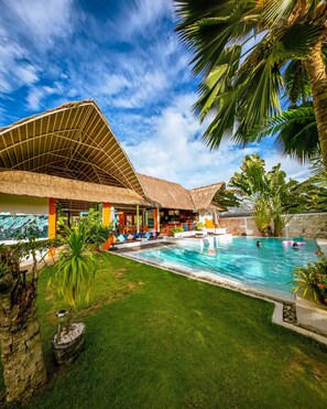 A pool area with a thatched-roof structure, a grassy lawn, and palm trees.