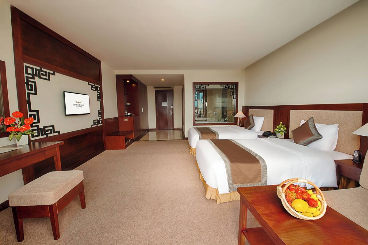 Habitación de hotel con dos camas, un escritorio, una silla, una mesita de noche con una cesta de frutas y un panel decorativo en la pared.