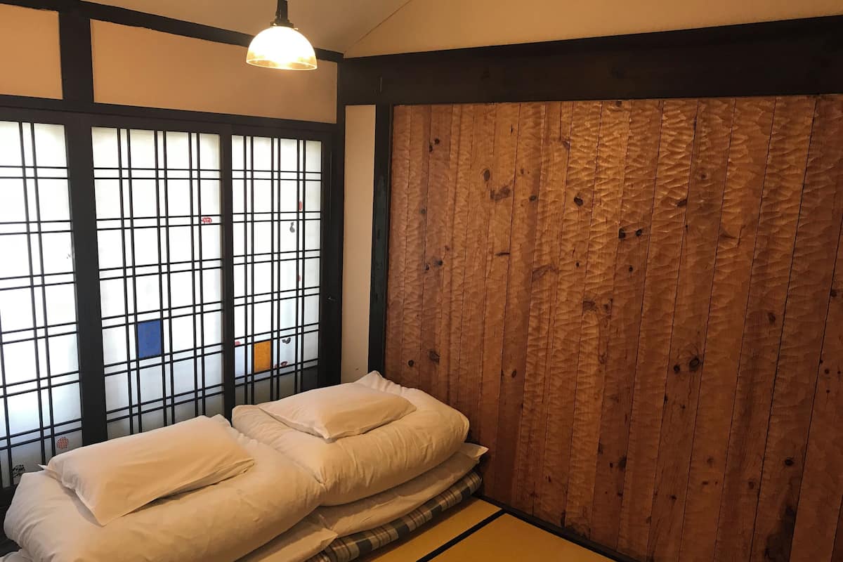 A room with tatami flooring, a low bed with white bedding, a shoji screen, and a hanging light fixture.