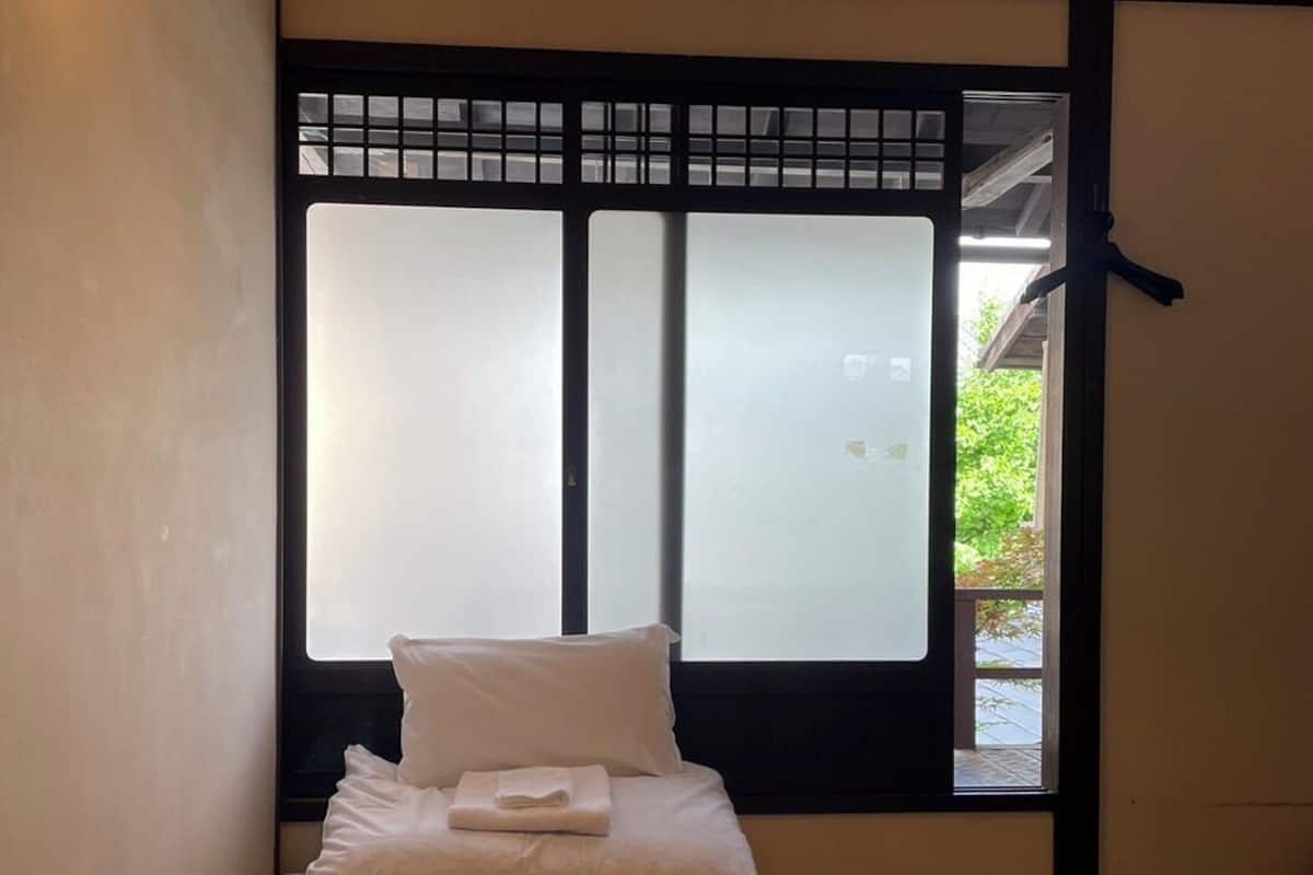 A room with tatami flooring, a bed with white bedding, a hanging light fixture, and a window with a view of greenery.