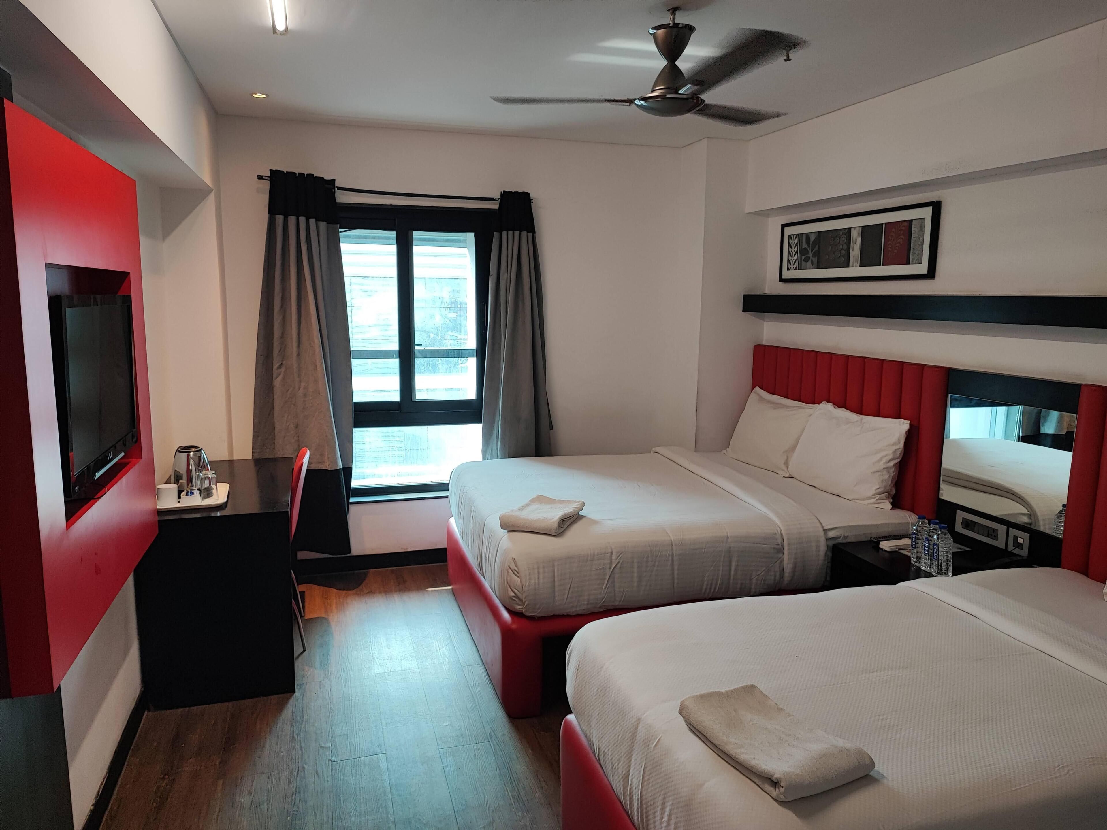 A hotel room with two beds, a TV, a desk with a coffee maker, a ceiling fan, and a window with curtains.
