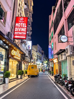 A street view of Hotel Divine Destination with a prominent red sign, a yellow van, and parked motorcycles.