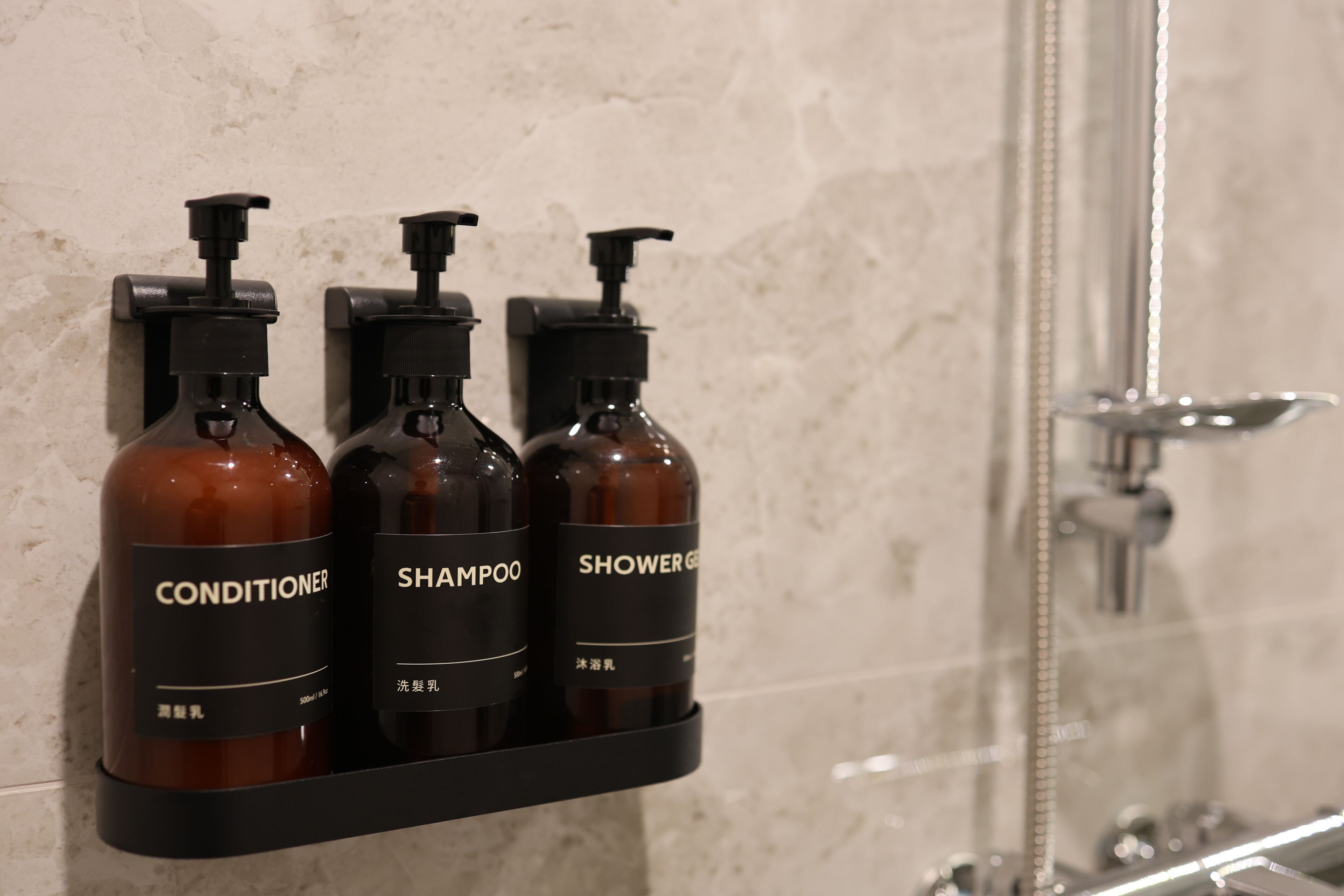 A bathroom with a shower, a mirror, and three bottles of toiletries on a shelf.