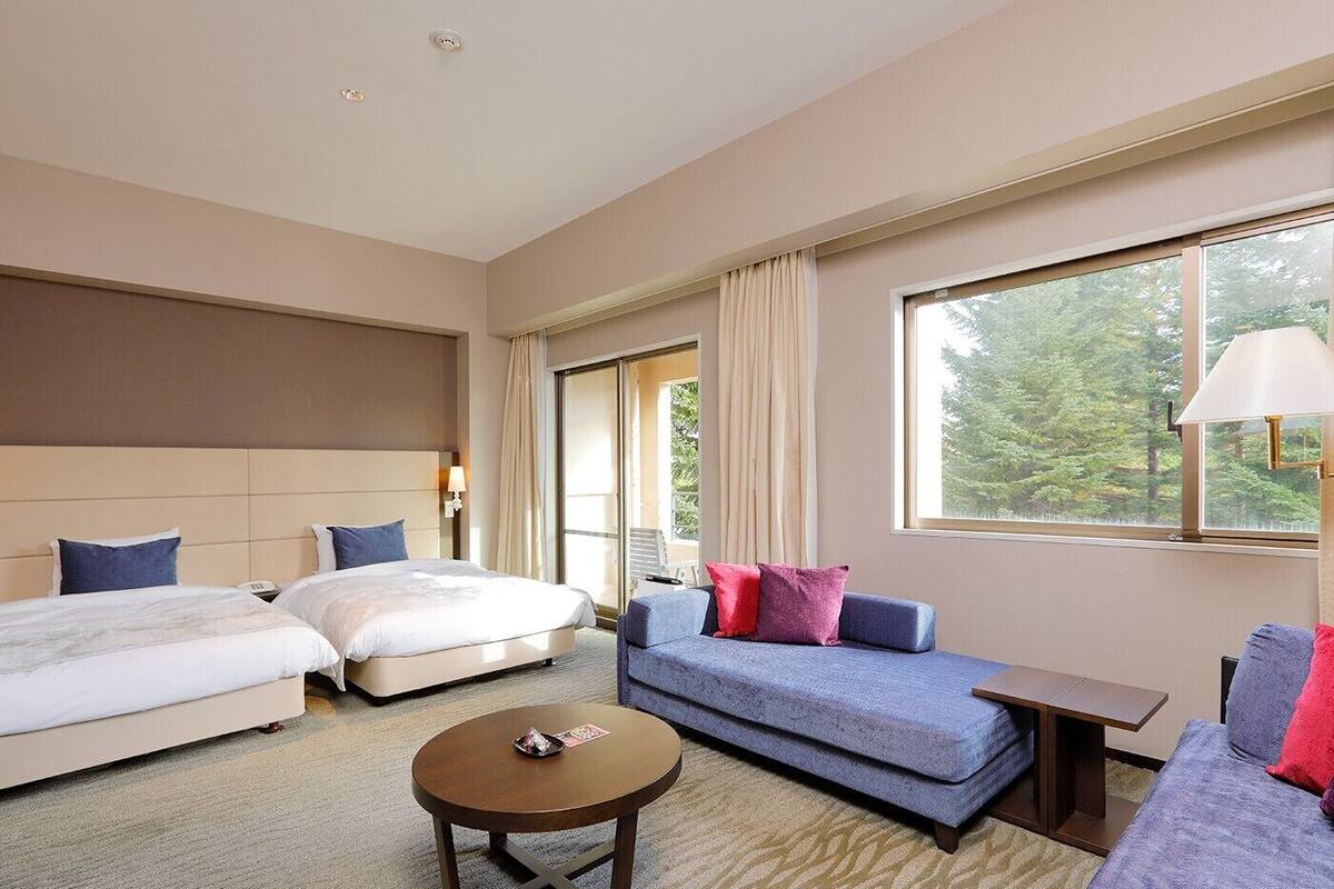 Habitación de hotel con dos camas, un sofá, una mesa de centro y una ventana con vistas a árboles.
