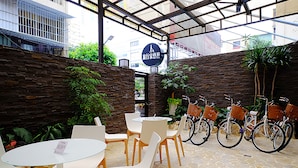 A hotel courtyard with stone walls, white chairs, and tables. There are bicycles parked and a sign indicating the hotel's location.