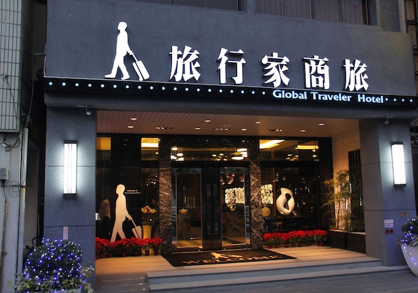 Exterior do Hotel Global Traveler, com letreiros iluminados, entrada convidativa e plantas ornamentais em vasos.