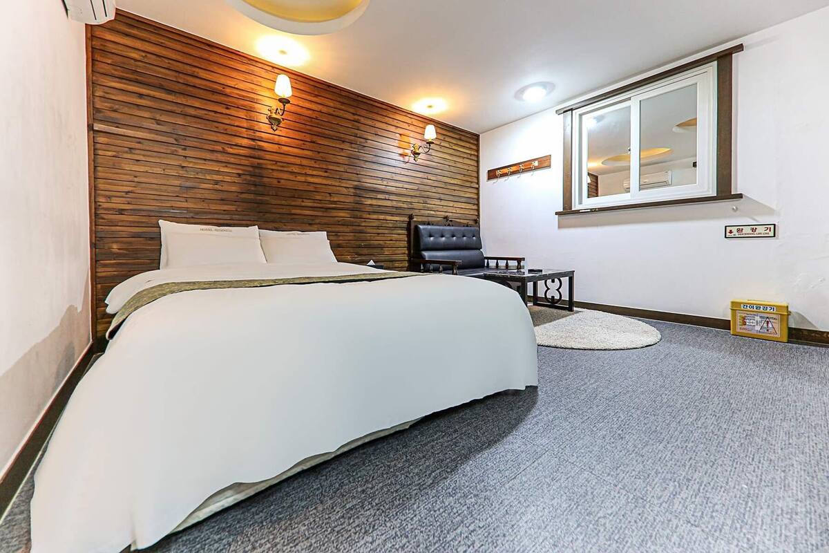 A hotel room with a large bed, a wooden headboard, two wall sconces, a small table, a chair, a round rug, and a window with a view.