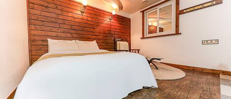 Quarto de hotel com cabeceira de madeira, cama com roupa de cama branca, uma mesinha, uma cadeira e um espelho na parede.