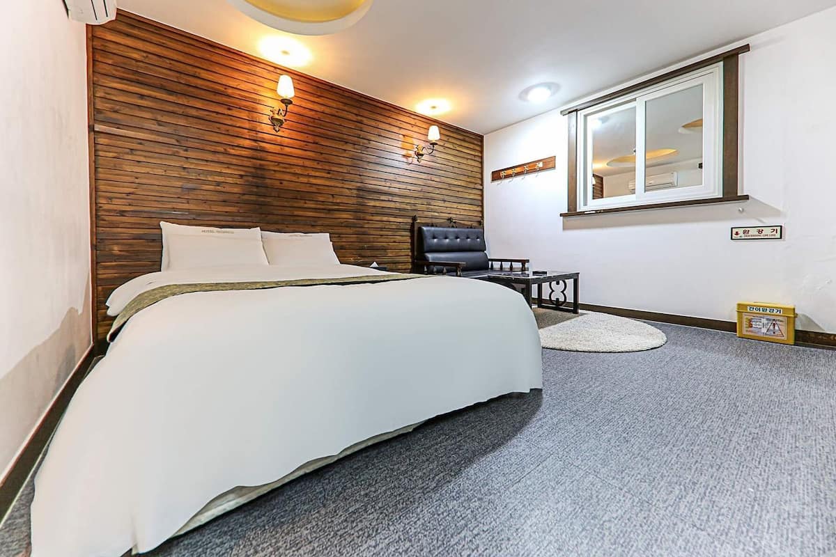 A hotel room with a large bed, a wooden headboard, two wall-mounted lights, a small table, a chair, a round rug, and a window with a view.