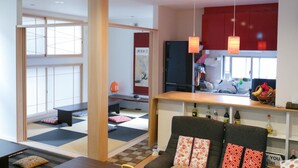 Una sala moderna con esteras de tatami, un sofá con cojines estampados, una zona de cocina con un refrigerador y una mesa de comedor con una cesta de fruta.