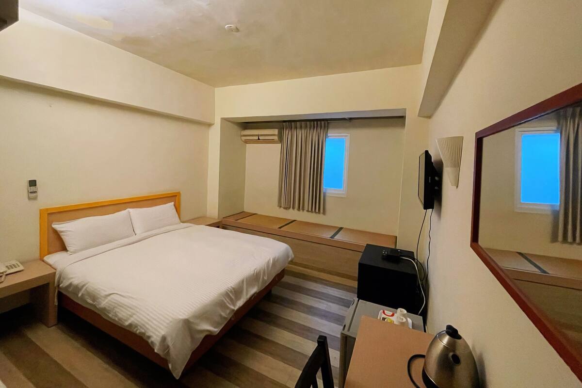 A hotel room with a single bed, a desk with a kettle, a flat-screen TV, and a window with curtains.