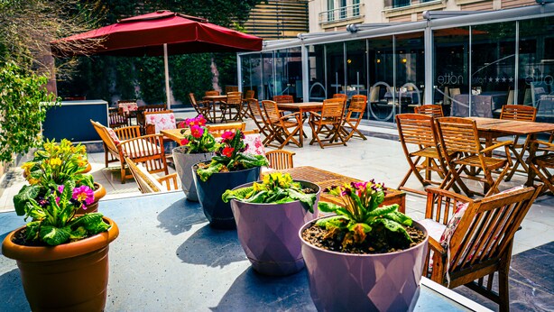 Un cafè a l'exterior amb taules i cadires de fusta, plantes en testos i un parasol vermell.