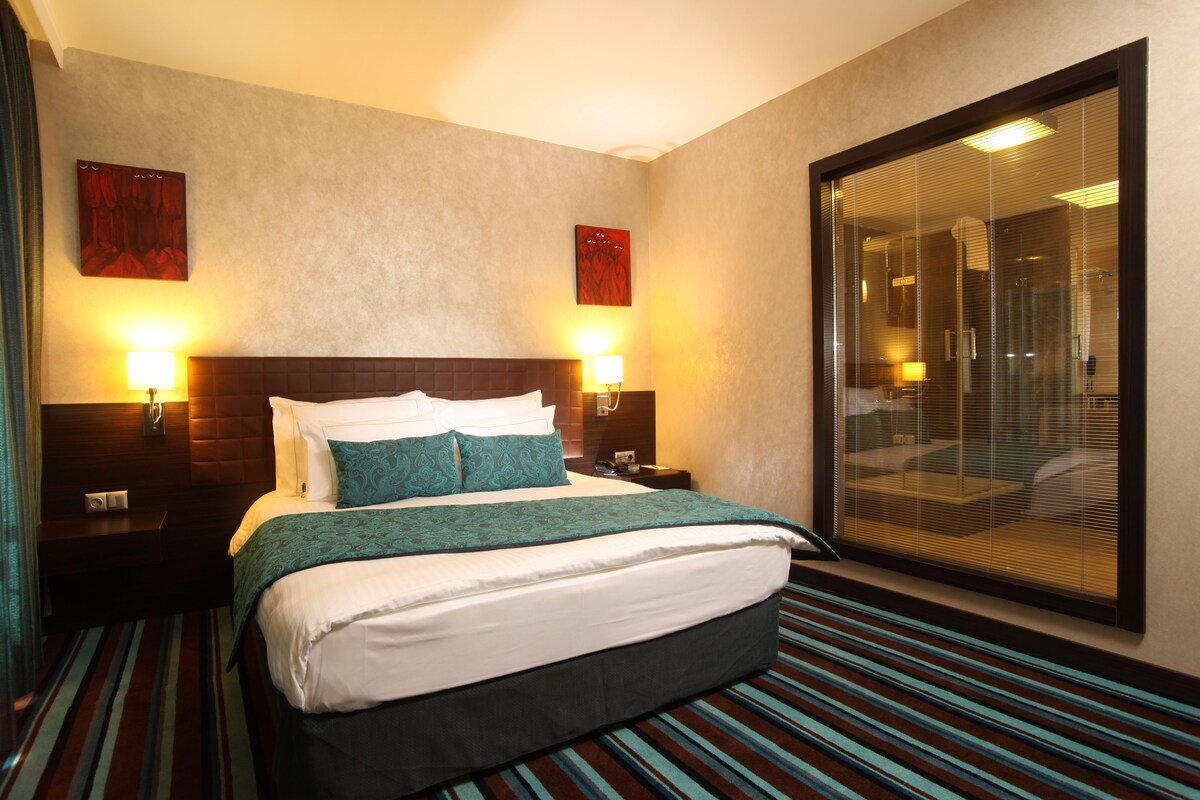 Habitación de hotel con una cama grande, dos lámparas de noche, una ventana con persianas y una obra de arte colgada en la pared.