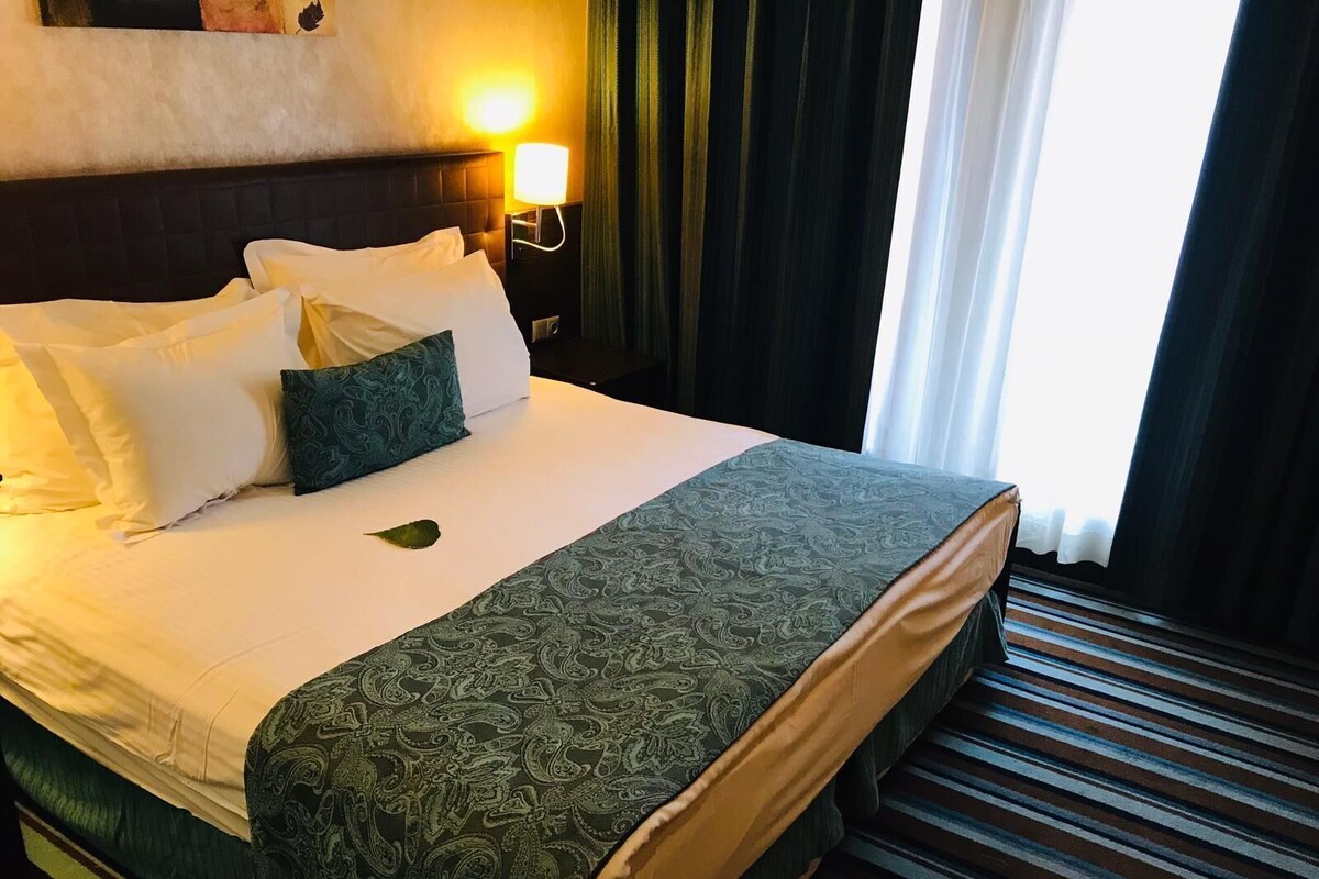 Una habitación de hotel con una cama, una mesita de noche, una lámpara y una ventana con cortinas.